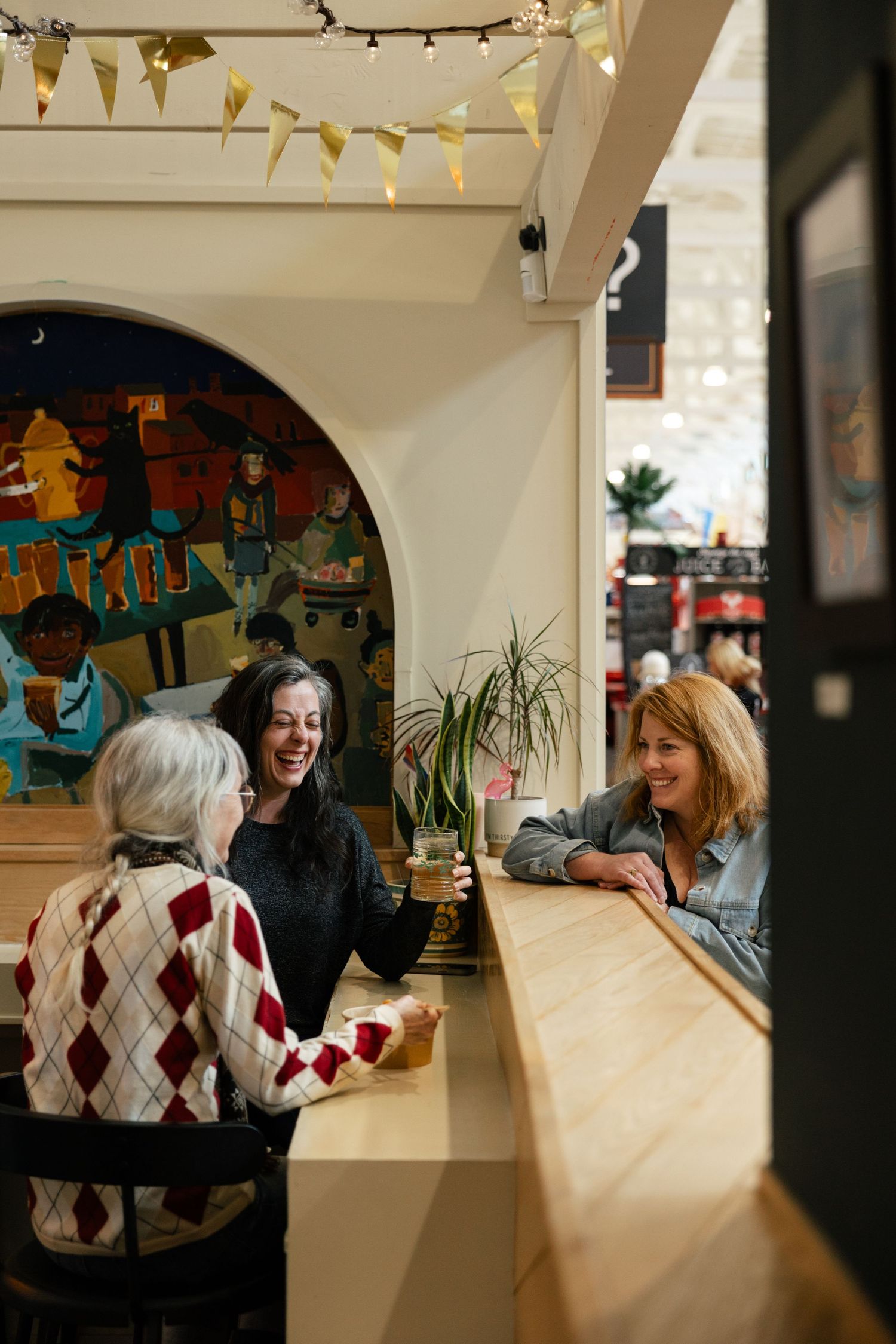 People chatting and laughing at a cafe counter with geometric patterns on the wall and festive bunting overhead.