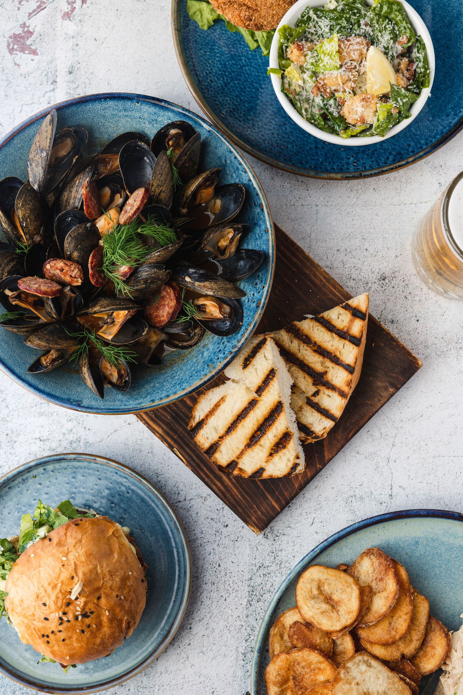 Overhead view of seafood appetizers including mussels, grilled bread, burger, and potato chips on blue ceramic plates.