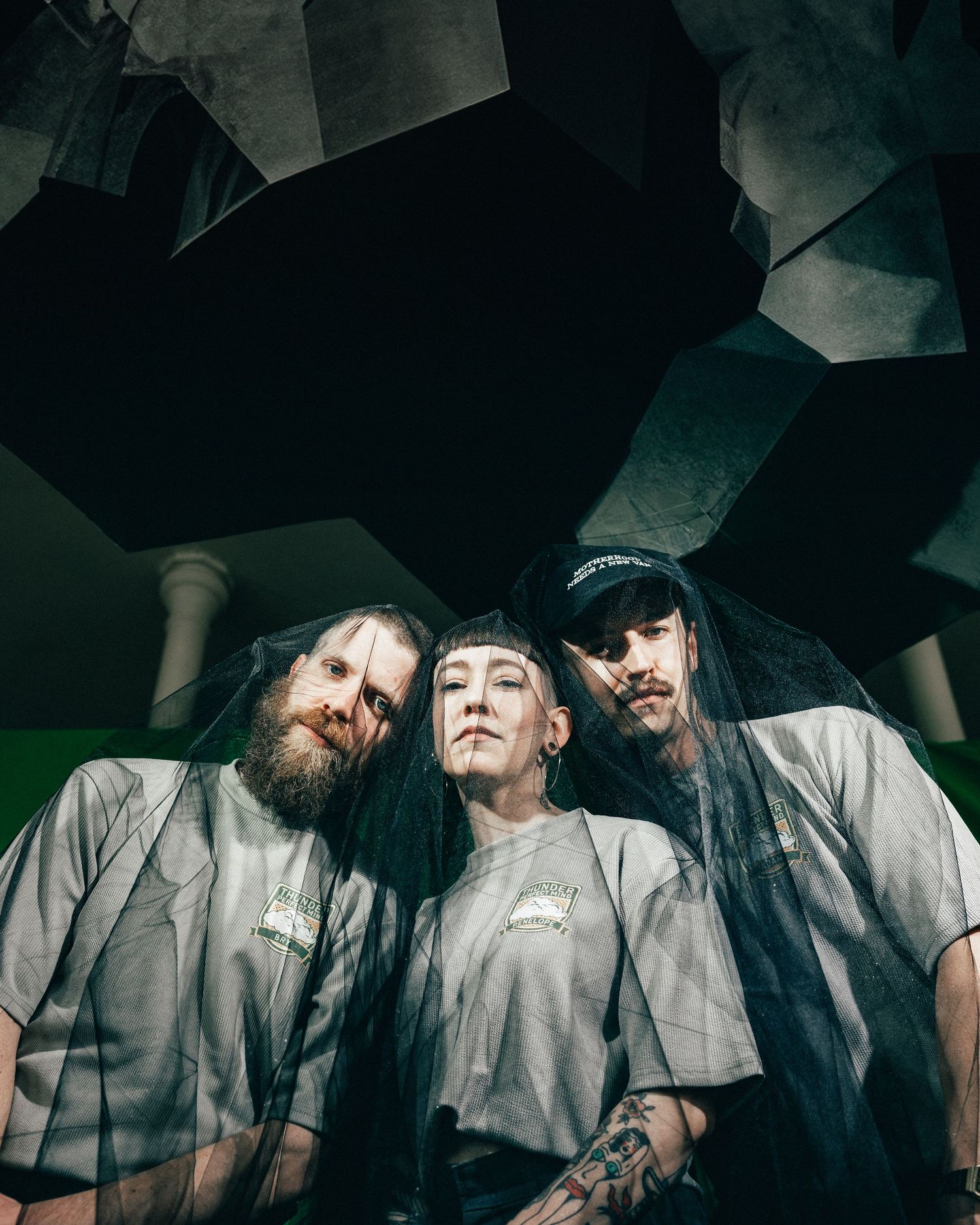 Artistic low-angle portrait of three bandmates wearing matching light colored shirts against a dark geometric background.