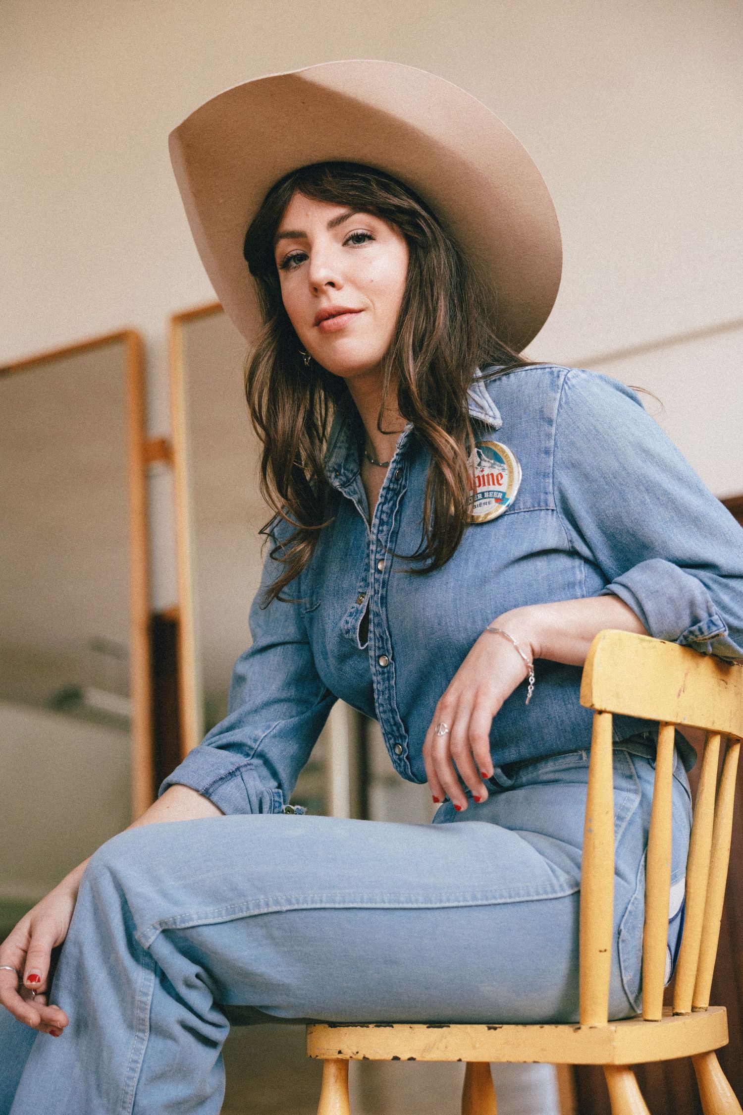 Style portrait featuring denim jumpsuit and beige wide-brimmed hat while seated on wooden chair in natural light.