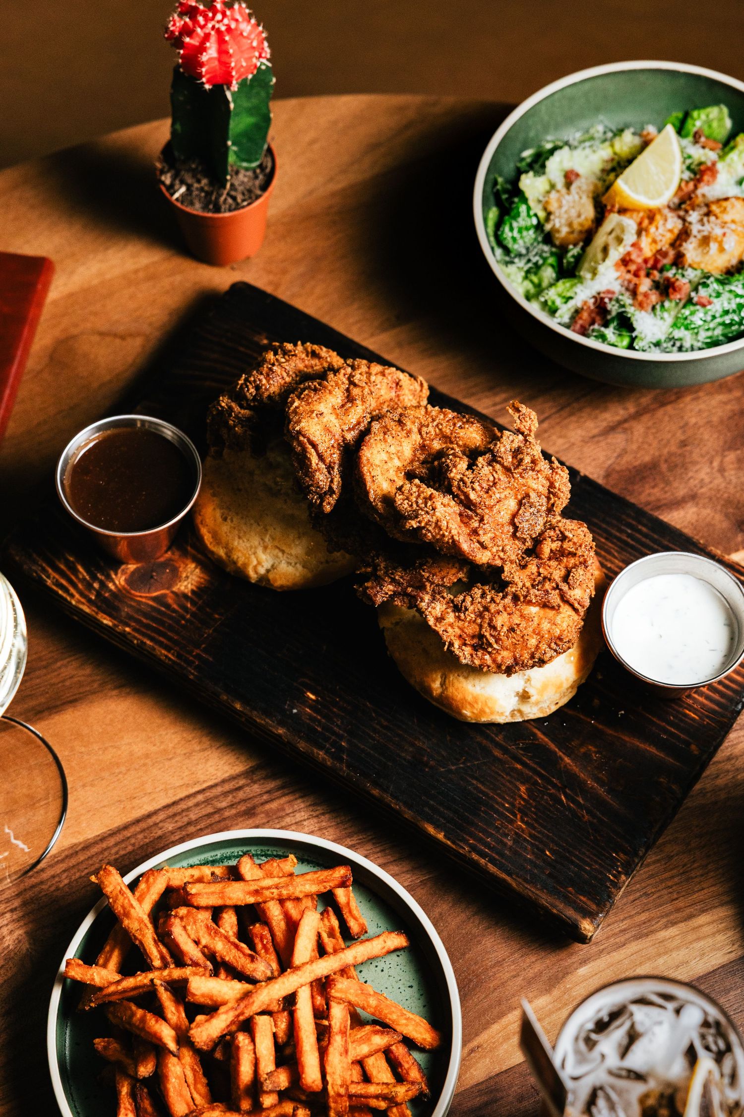 A rustic wooden board with fried chicken sandwiches served alongside sweet potato fries and salad in a casual dining setup.