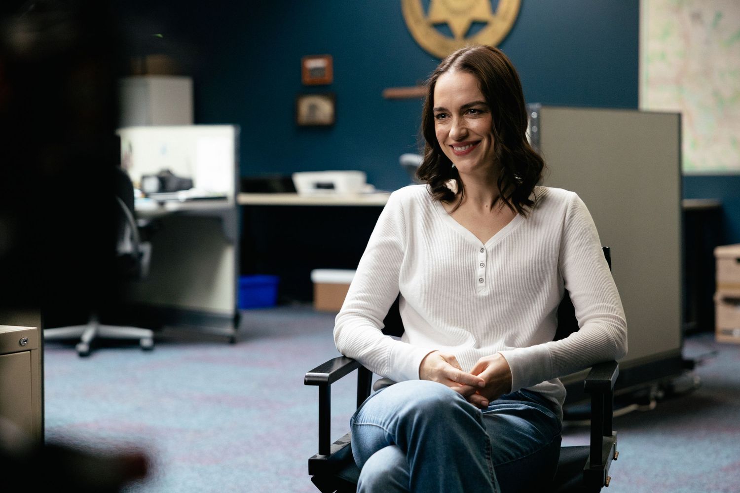 A person in a white henley shirt sits in an office chair against a dark teal wall.
