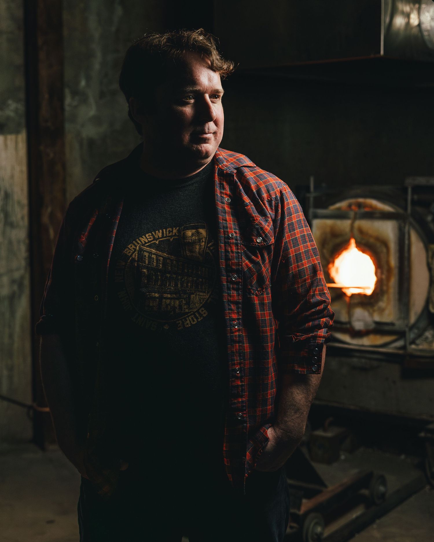 Person in red plaid flannel shirt and dark t-shirt standing in rustic interior with warm lighting in background.