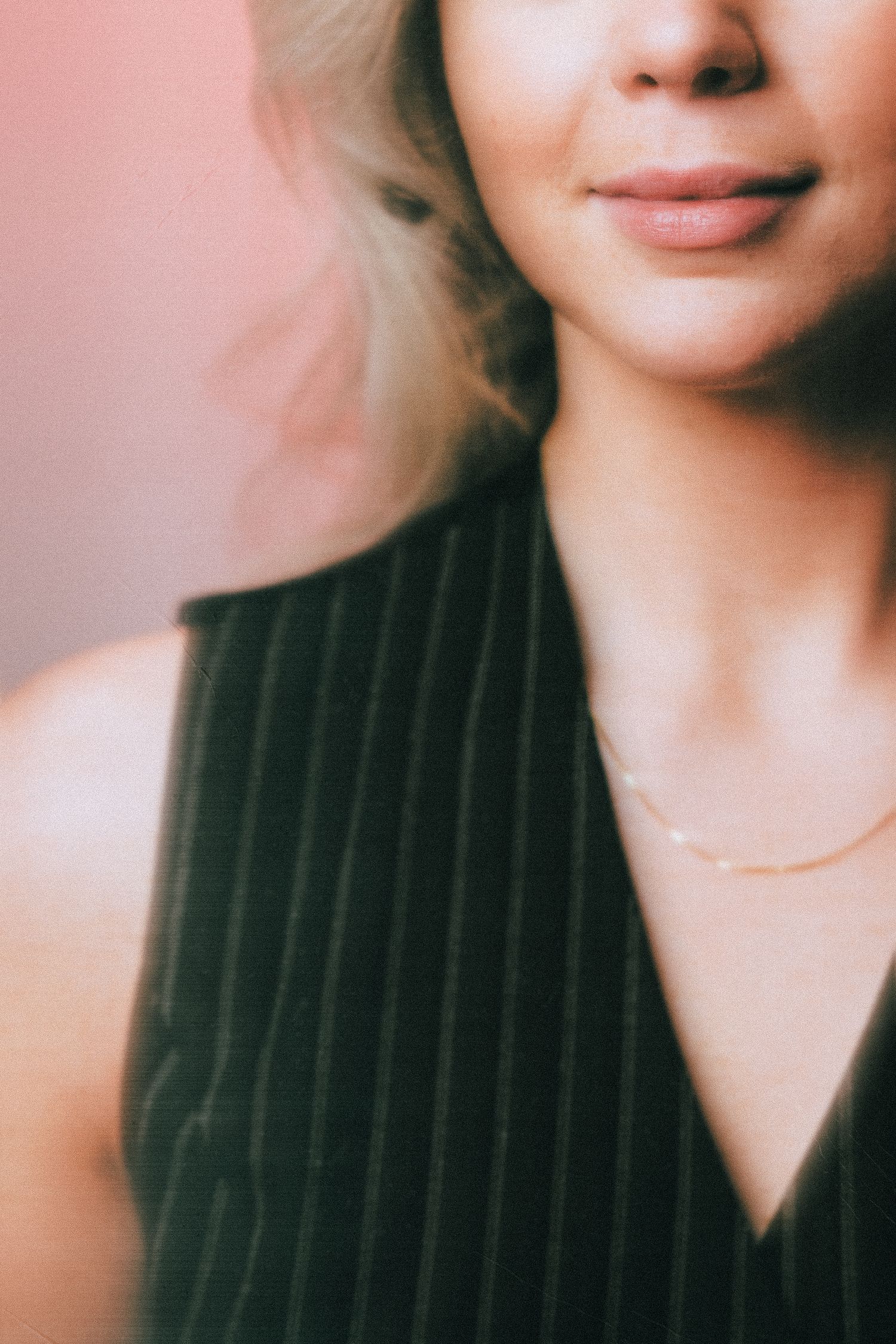 Cropped portrait showing lips and neckline in a pinstriped black top with delicate gold necklace against soft pink background.
