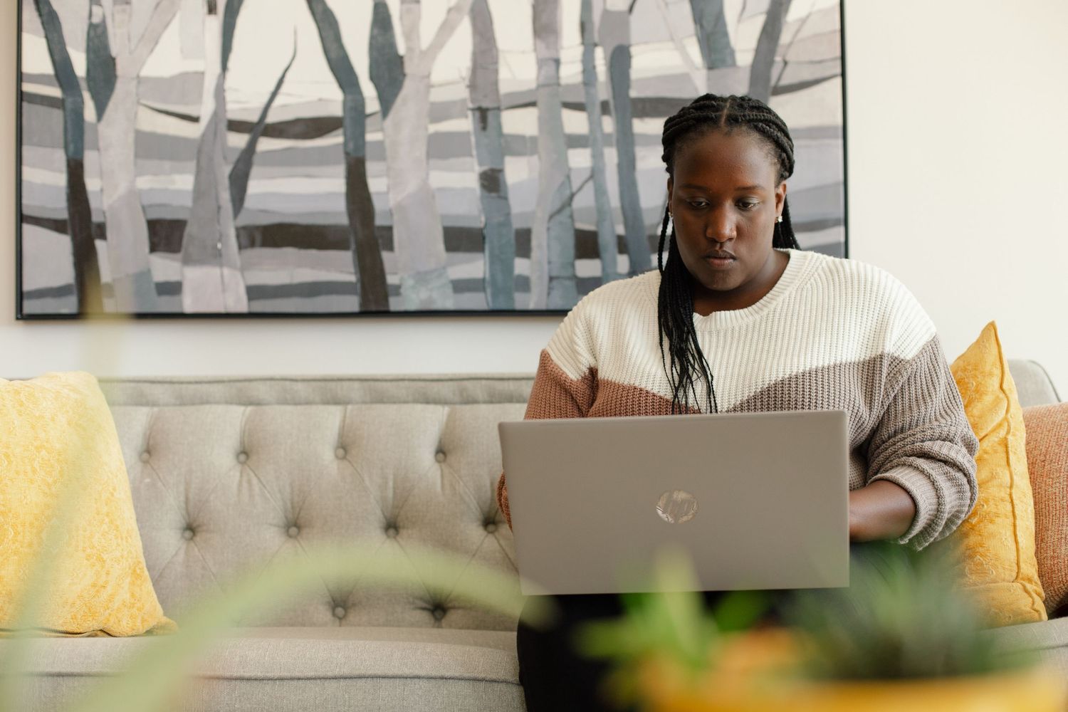 A person sits on a gray couch working on a laptop with abstract wall art in the background.
