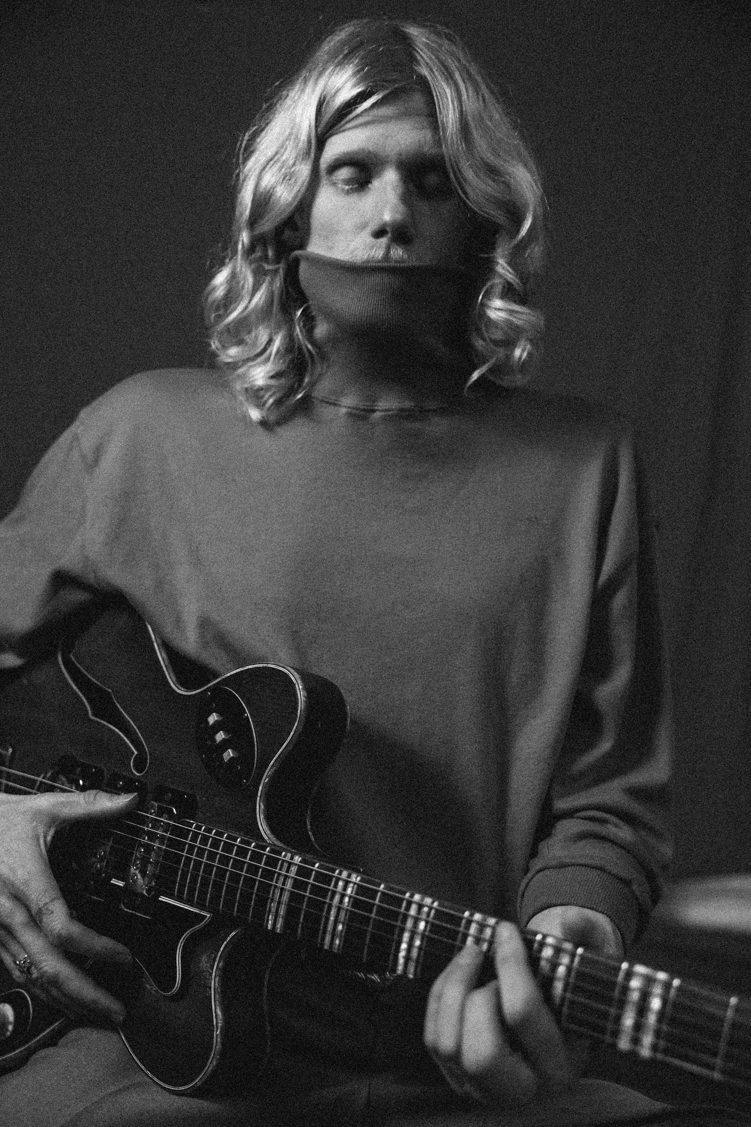Black and white portrait of a musician with blonde wavy hair holding an electric guitar, wearing a casual sweater.