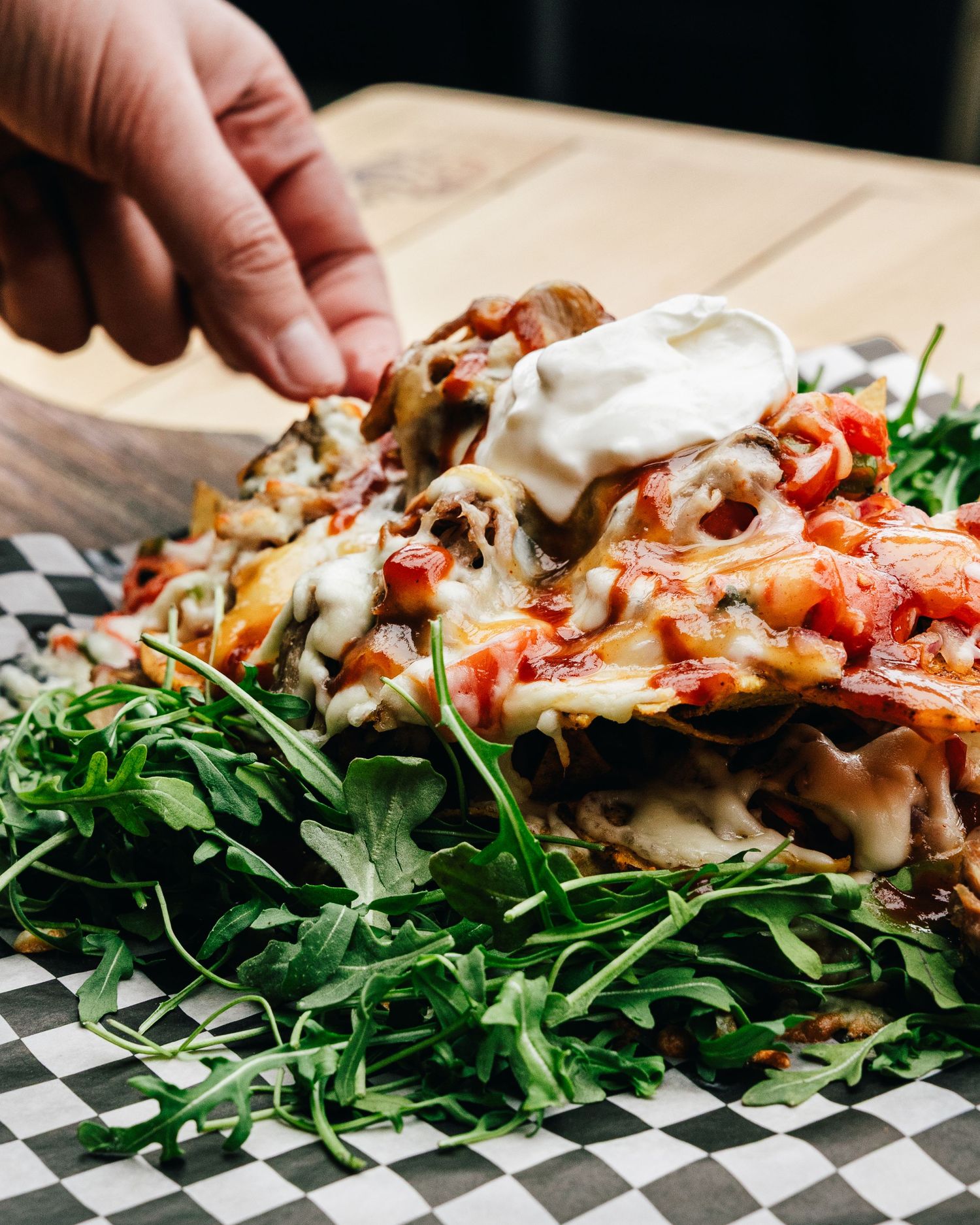 Loaded nachos topped with melted cheese, tomatoes, sour cream and fresh greens served on a checkered paper liner.
