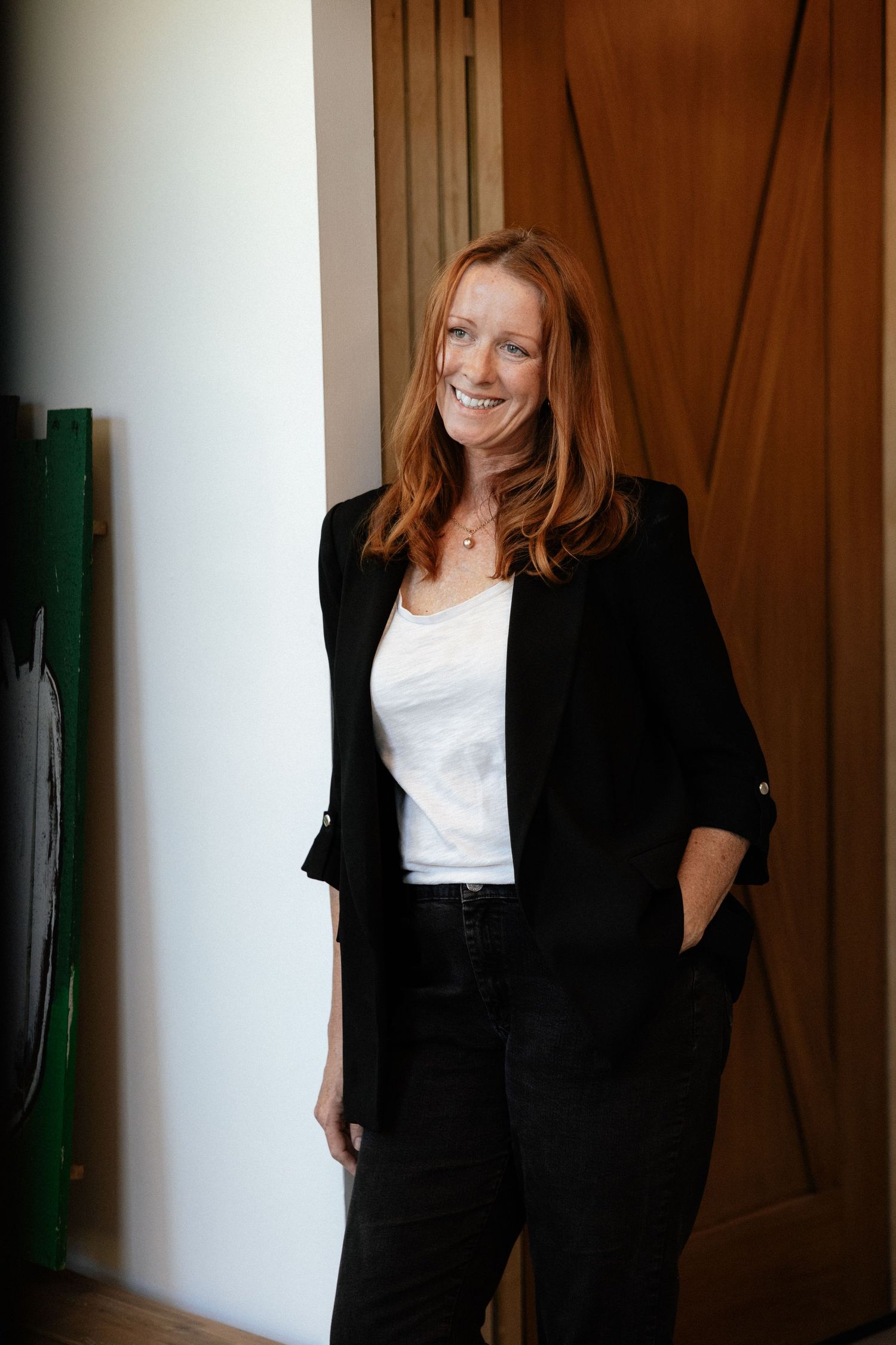 Professional portrait of someone with long red hair wearing a black blazer and white top standing against a wooden door.