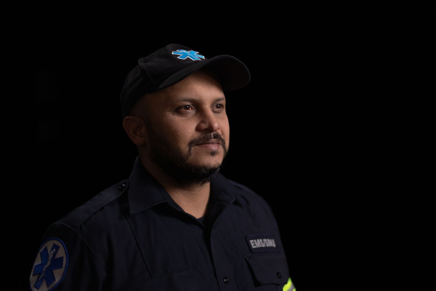 Professional headshot portrait of someone wearing a dark jacket and cap against a black background.