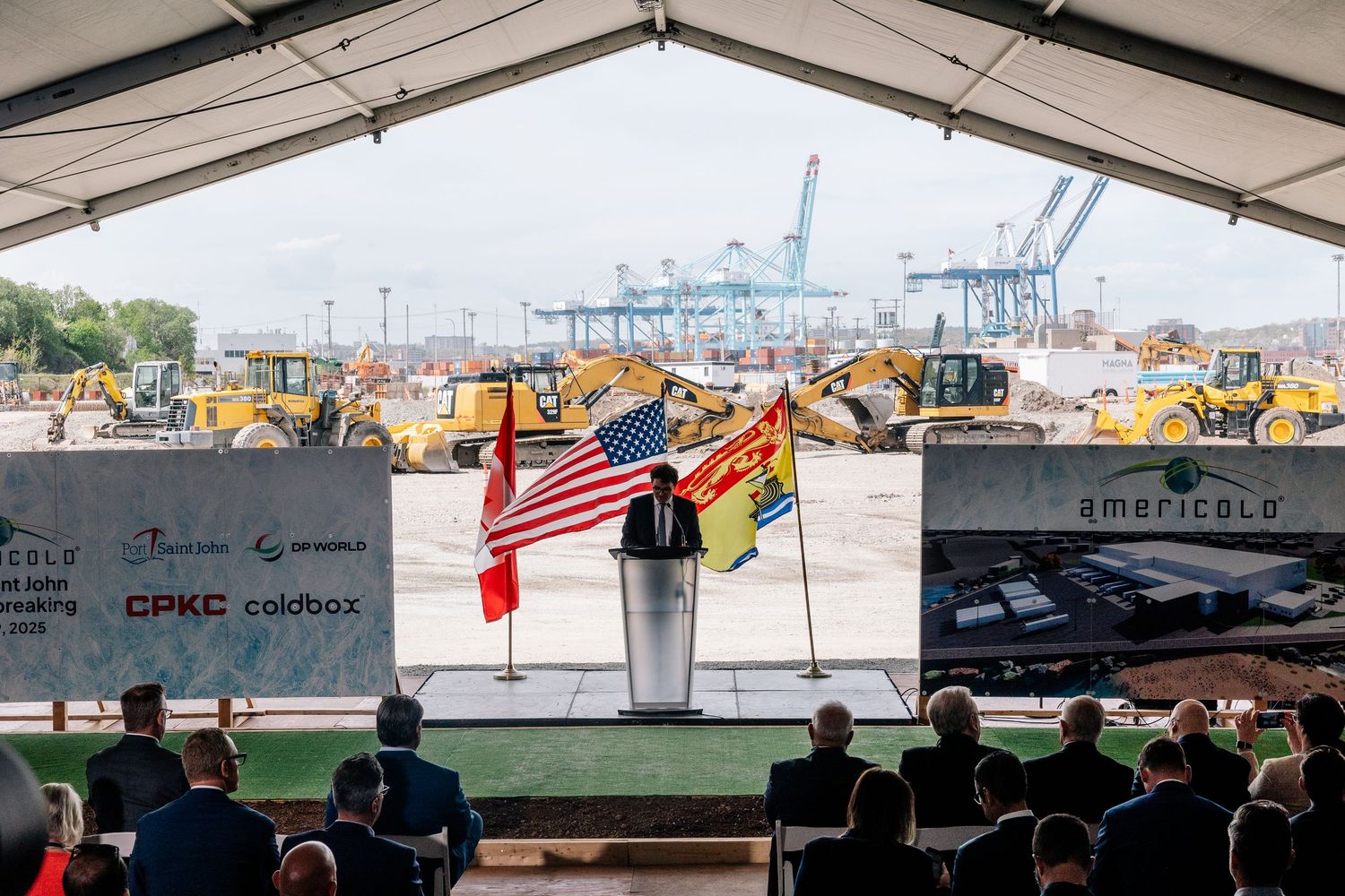Flags displayed at an outdoor ceremony with shipping container cranes visible in the background at an industrial port.
