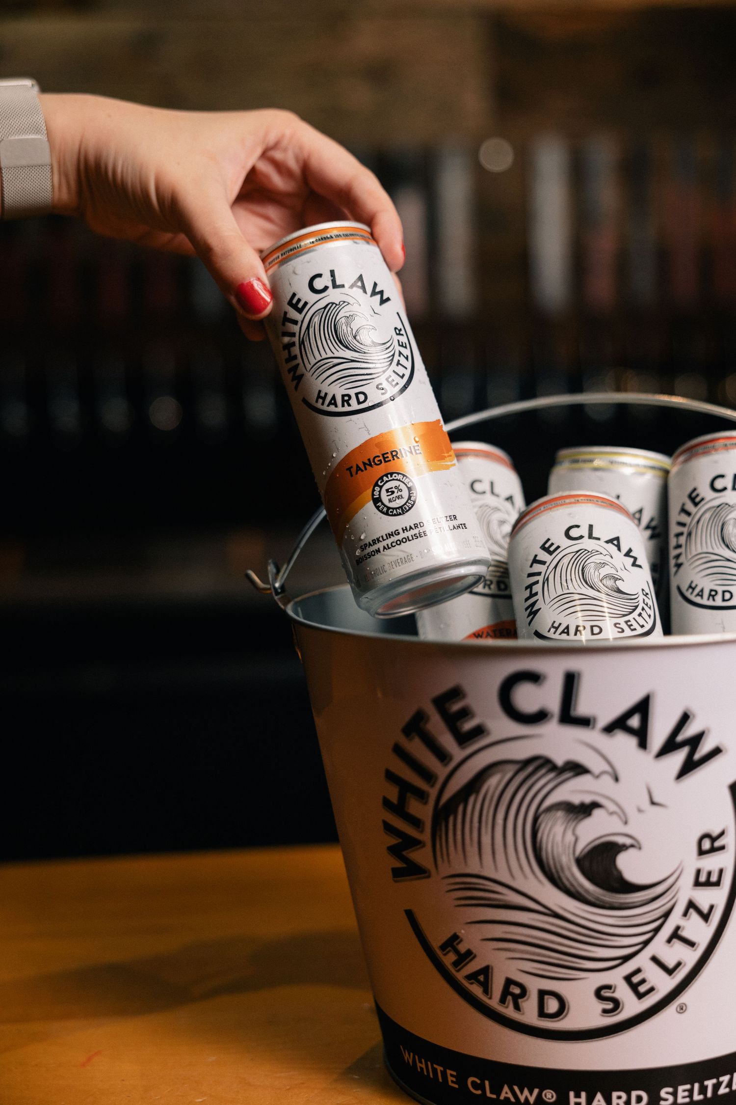 A hand placing a White Claw hard seltzer can into a branded ice bucket filled with other cans of the same drink.