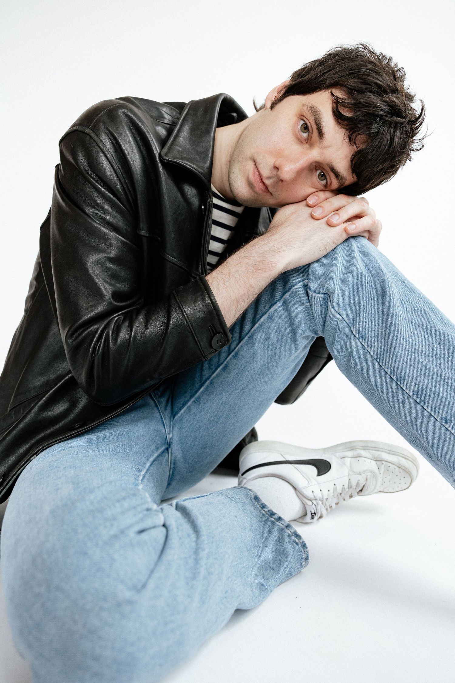 A person wearing a black leather jacket and striped shirt poses casually on a white background with light denim jeans.