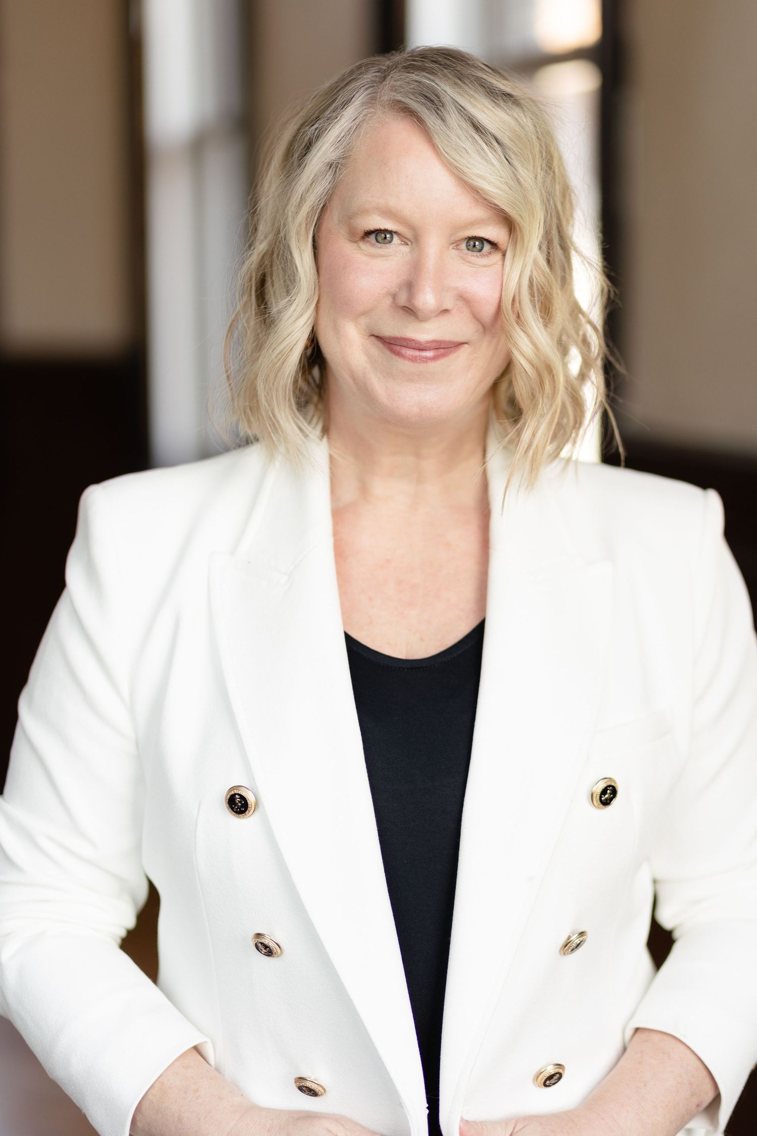 Professional headshot of someone wearing a crisp white blazer with gold buttons over a black top against a blurred background.