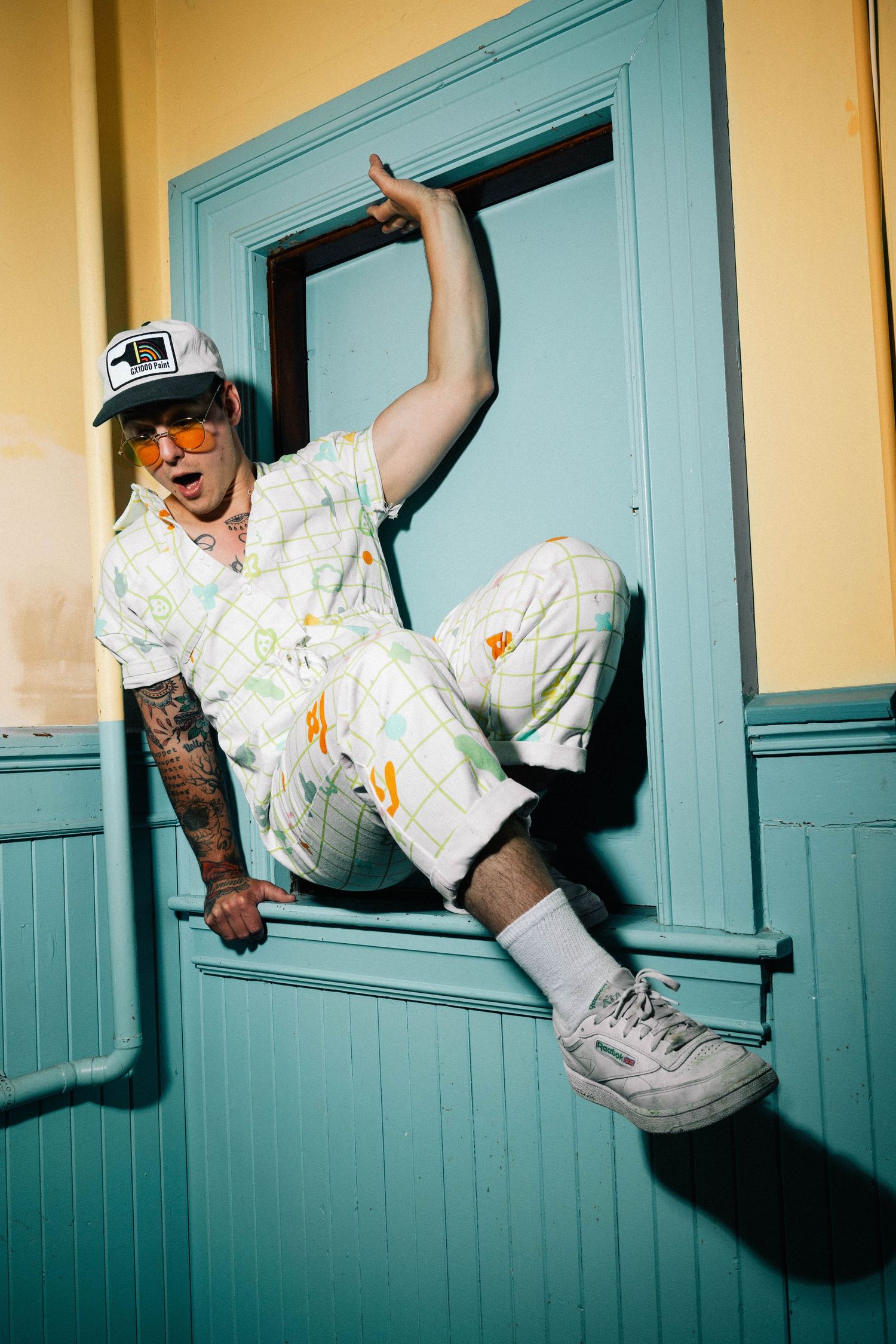 Person in retro sportswear and cap poses playfully in turquoise doorframe with dramatic flash lighting.