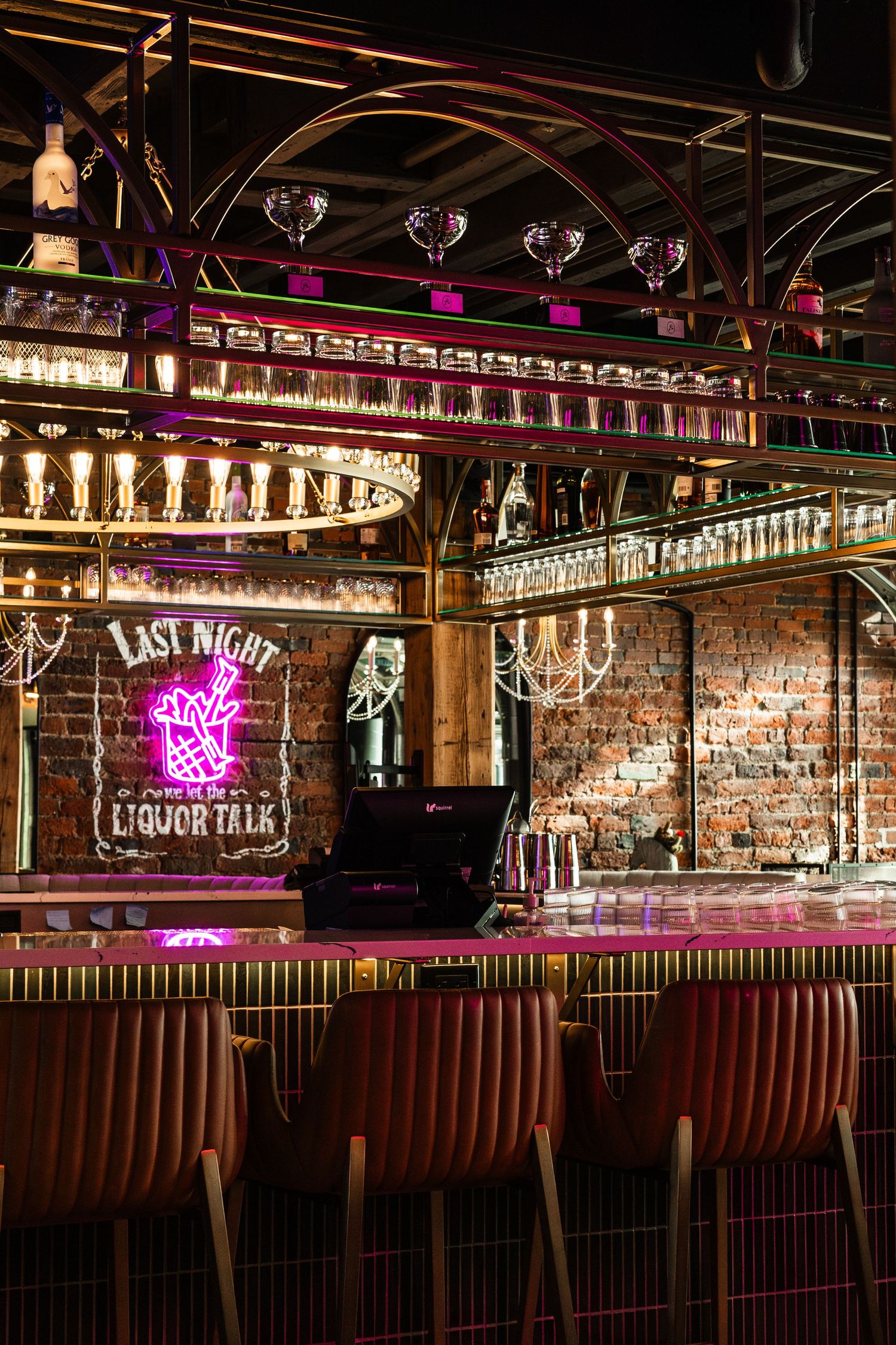 Stylish bar interior with neon 'Last Night Liquor Talk' sign, exposed brick walls and leather seating under crystal glassware.