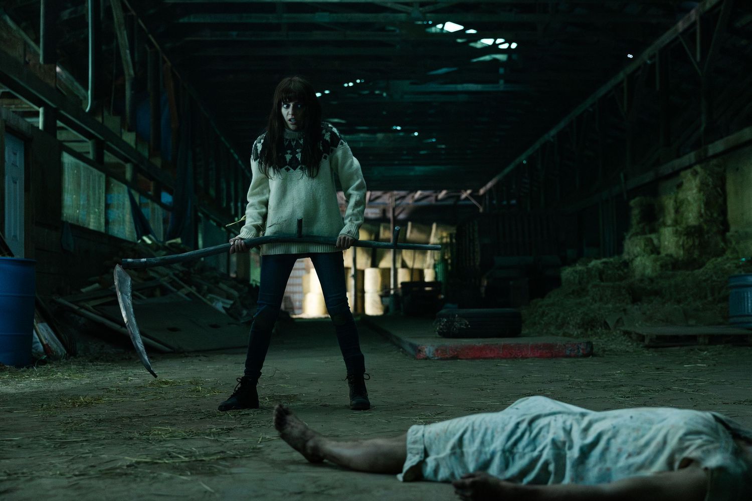 A dark and moody scene in an abandoned warehouse shows a person standing with a weapon near a body on the floor.