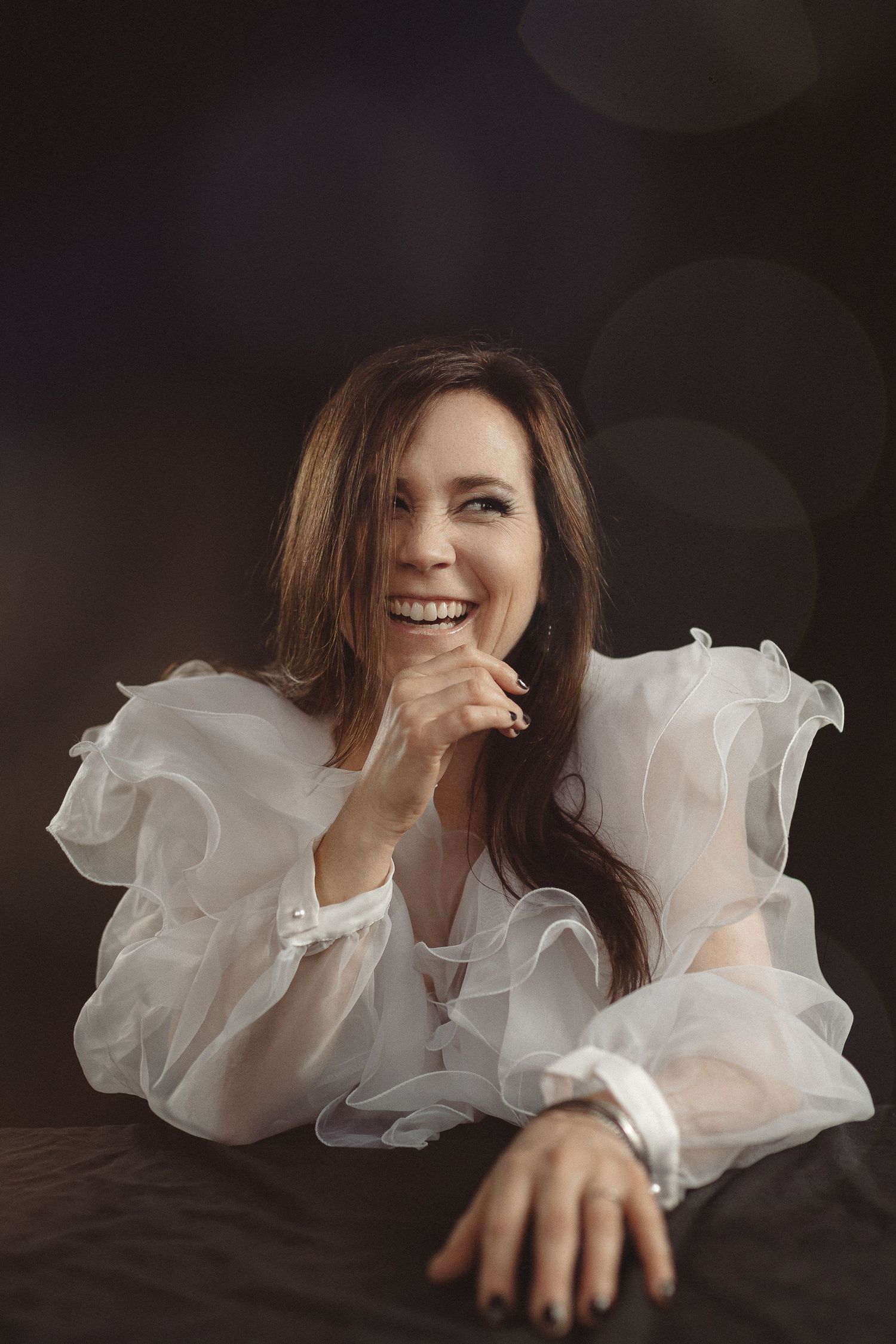 Portrait of someone wearing an elegant white ruffled dress while smiling warmly and resting their chin on their hand.