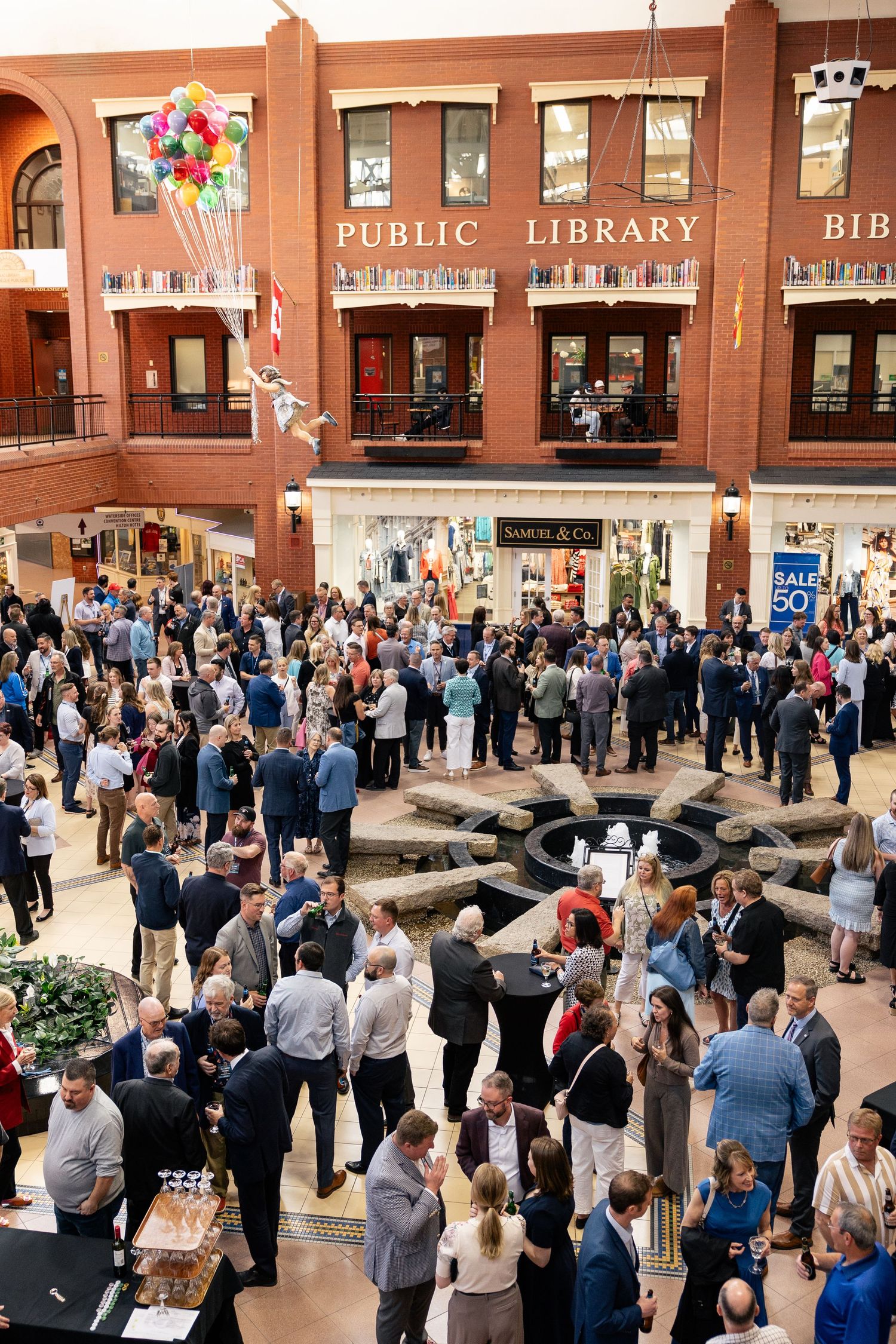 Large crowd gathering at an indoor public library event with festive balloons and people mingling in business attire.