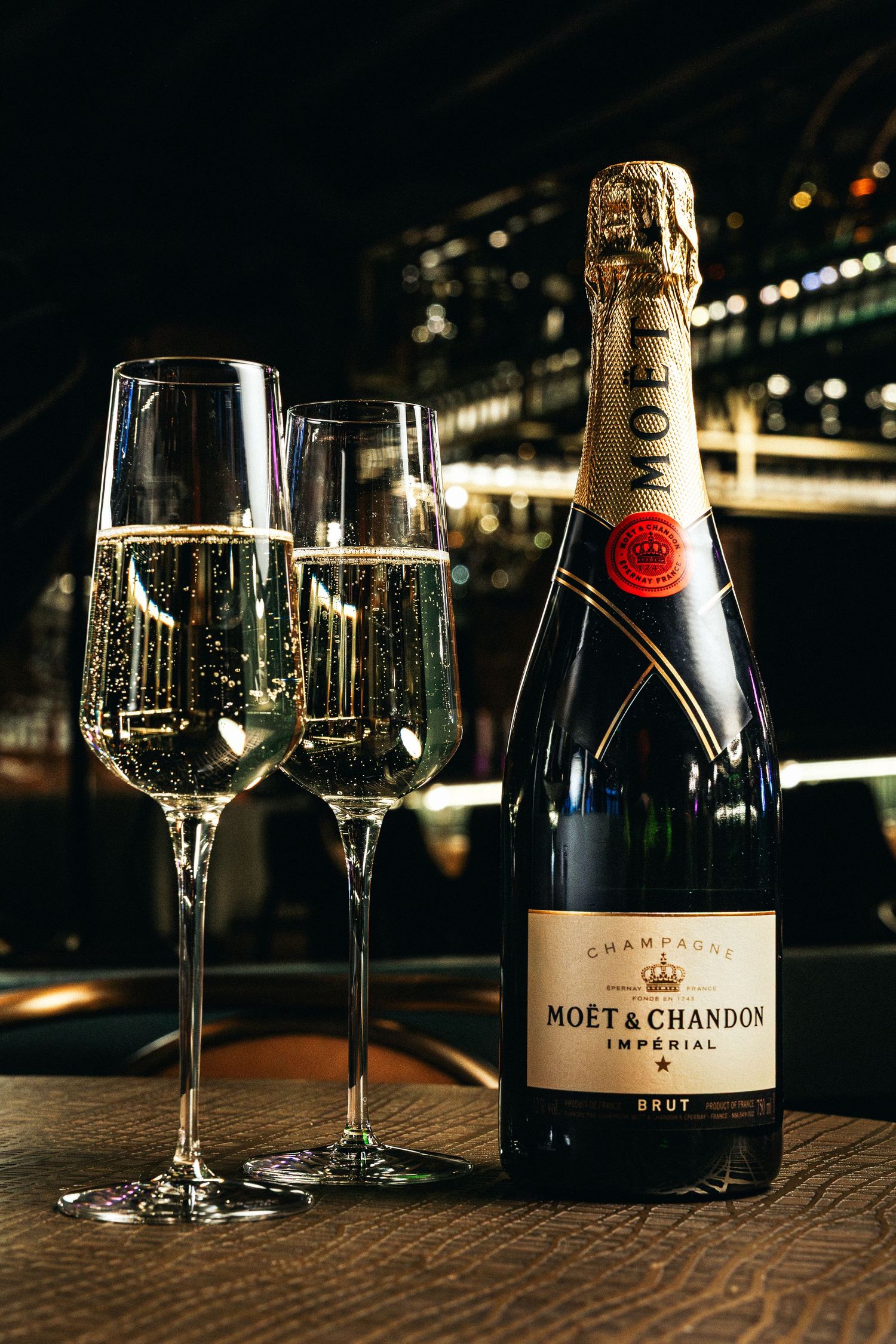 Moët & Chandon champagne bottle with two filled champagne flutes on a wooden surface with soft bokeh lights in background.