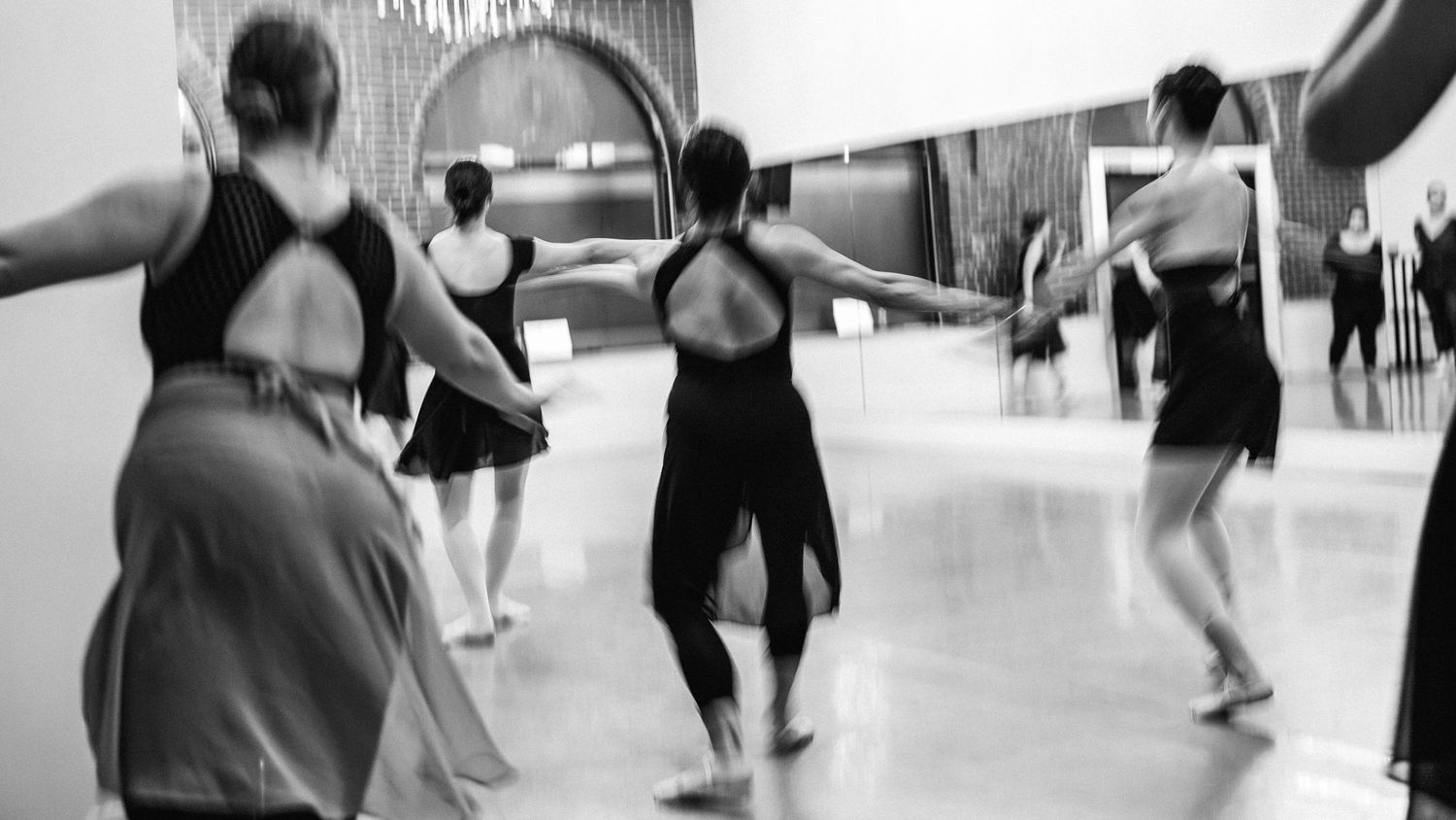Dancers in black leotards practicing ballet movements inside a dance studio hallway.