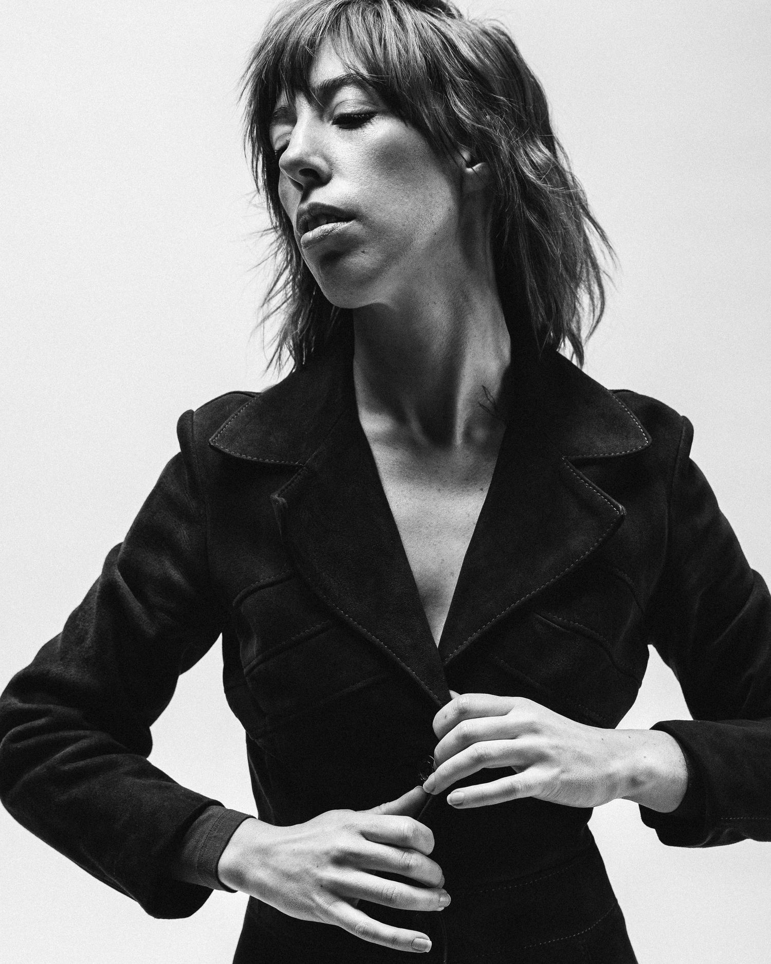 Black and white portrait of a figure in a fitted black blazer posing dramatically with closed eyes and windswept hair.