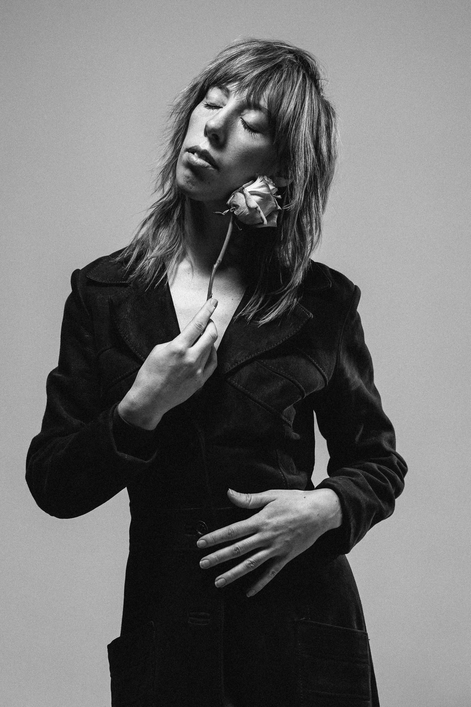Artistic black and white portrait of a person in a dark blazer holding a rose with eyes closed in contemplative pose.