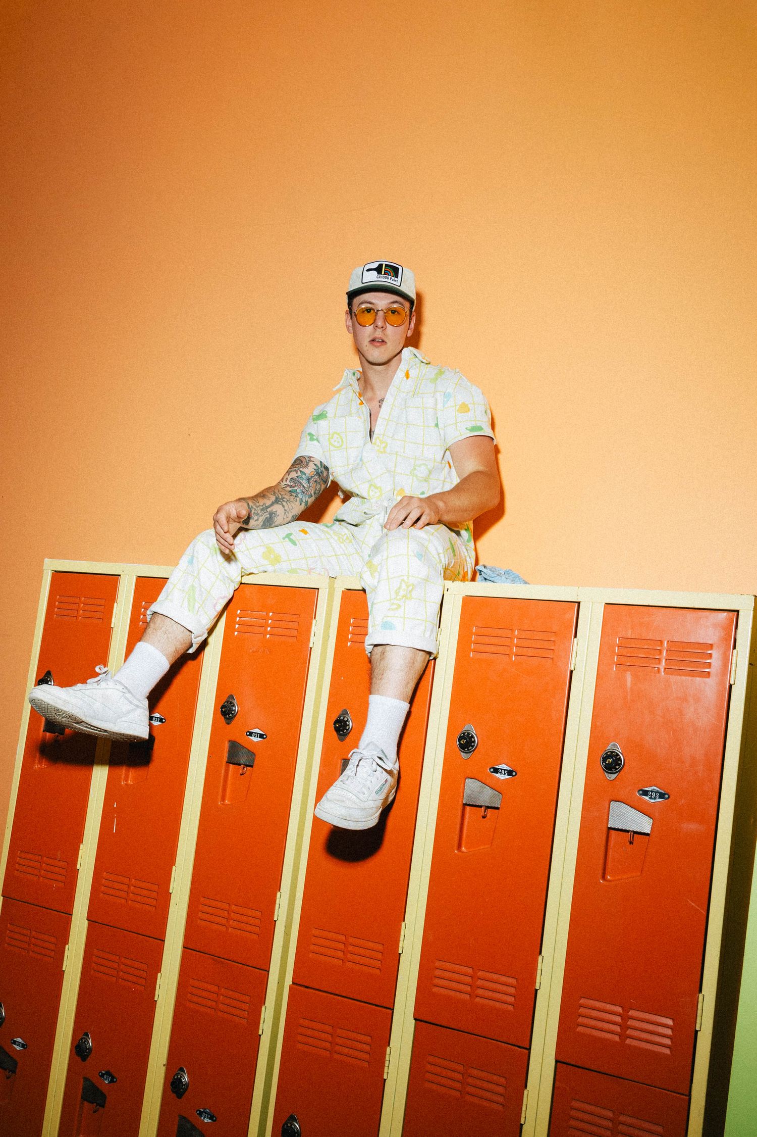 Person in white clothing and baseball cap sits atop orange lockers in stylized studio photograph.