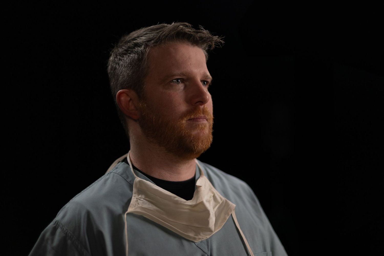 Person wearing a tan turtleneck and grey jacket looks to the side against a dark background.