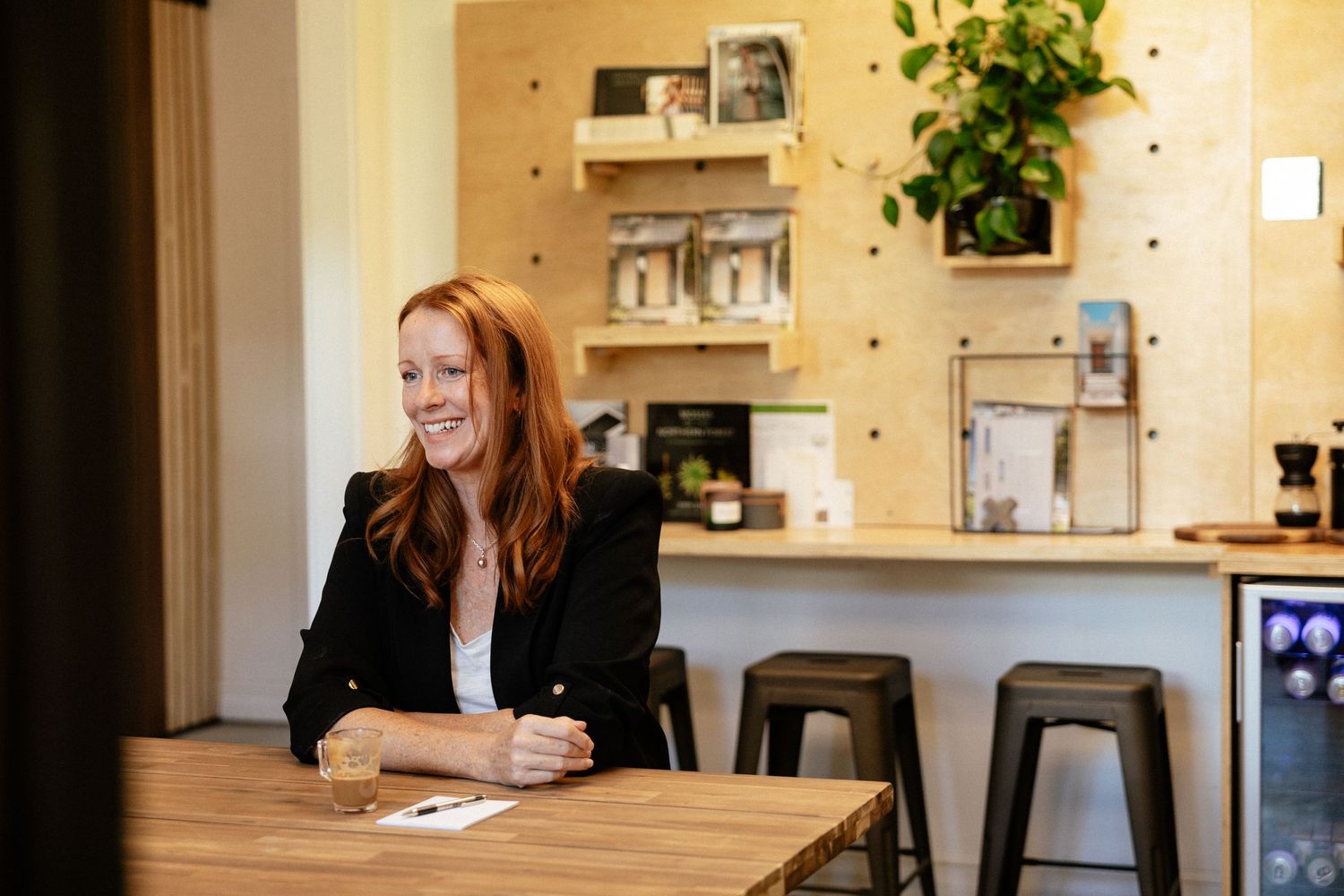 A person in a black blazer sits at a wooden table in a bright modern office space with shelving and plants.