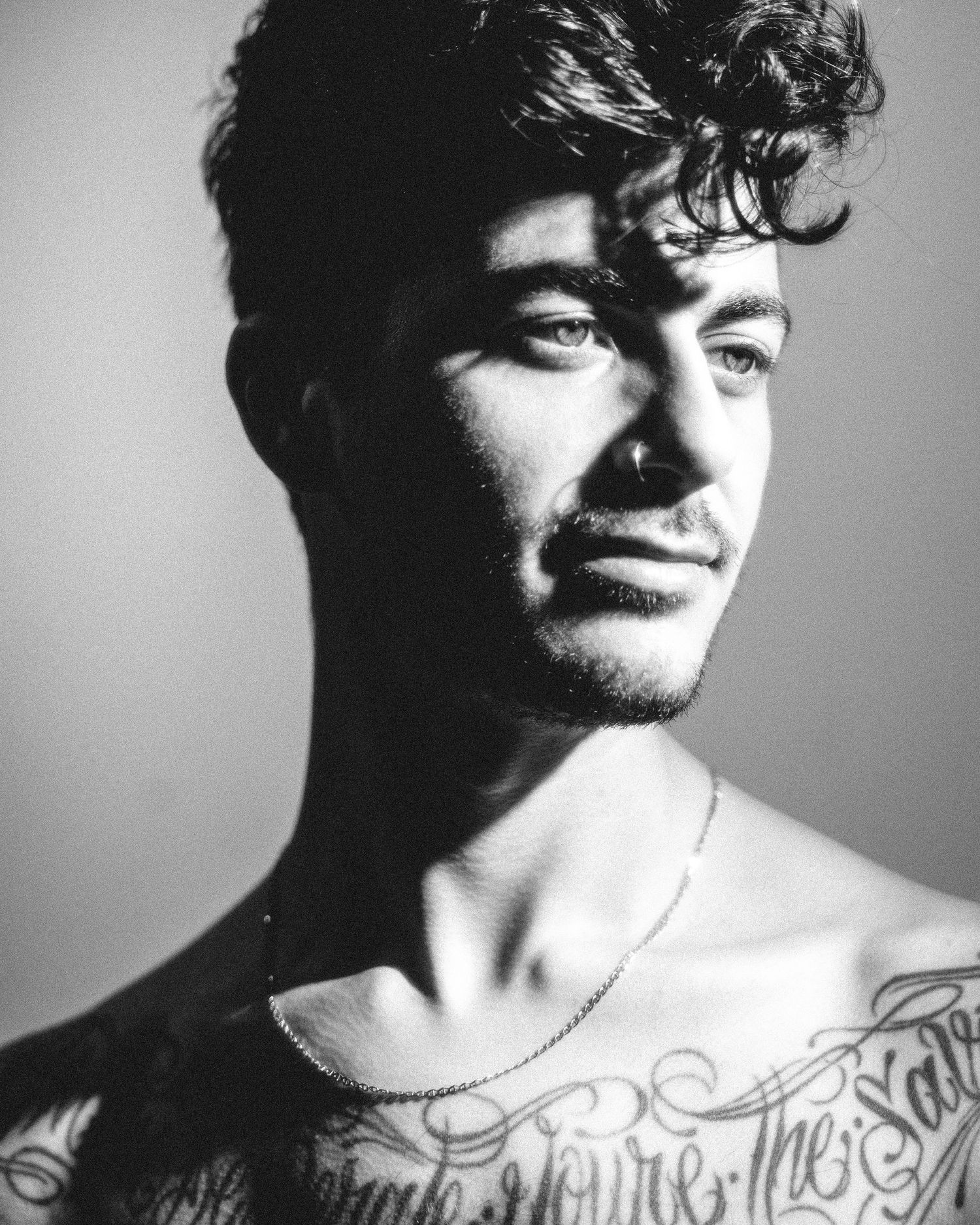 Black and white portrait showing torso with visible chest tattoos and necklace against dramatic studio lighting.