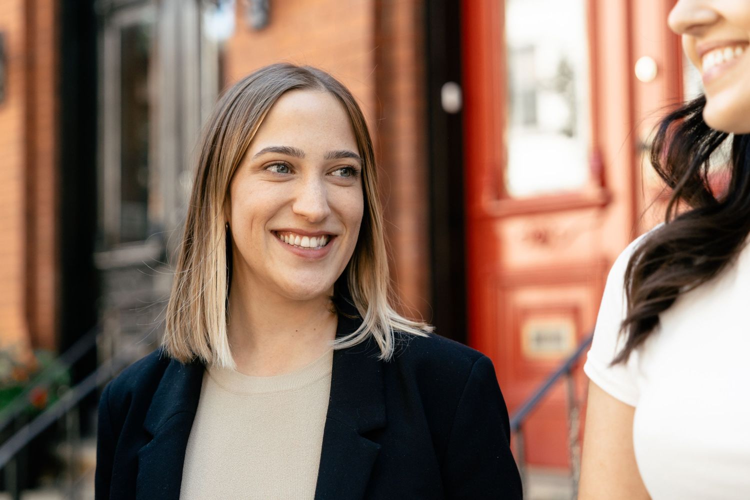 A collage of smiling individuals in front of red and white buildings, captured in a casual outdoor setting.