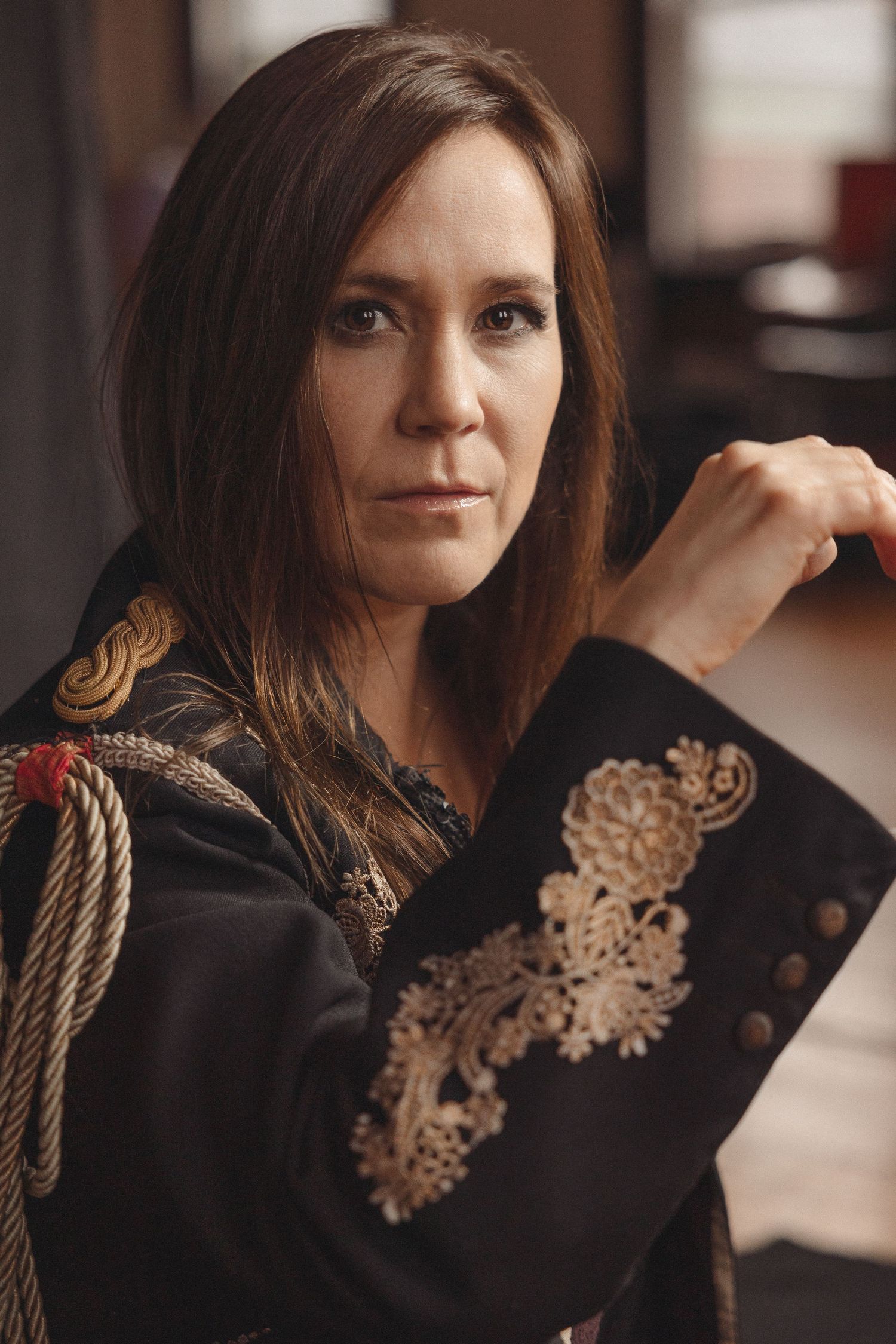A woman in a black embroidered top poses for a dramatic portrait with long brown hair.