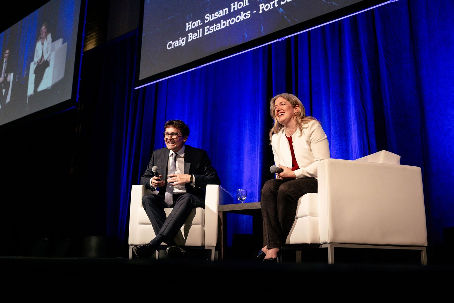 Two speakers sit in white chairs on stage with blue curtain backdrop during panel discussion event.