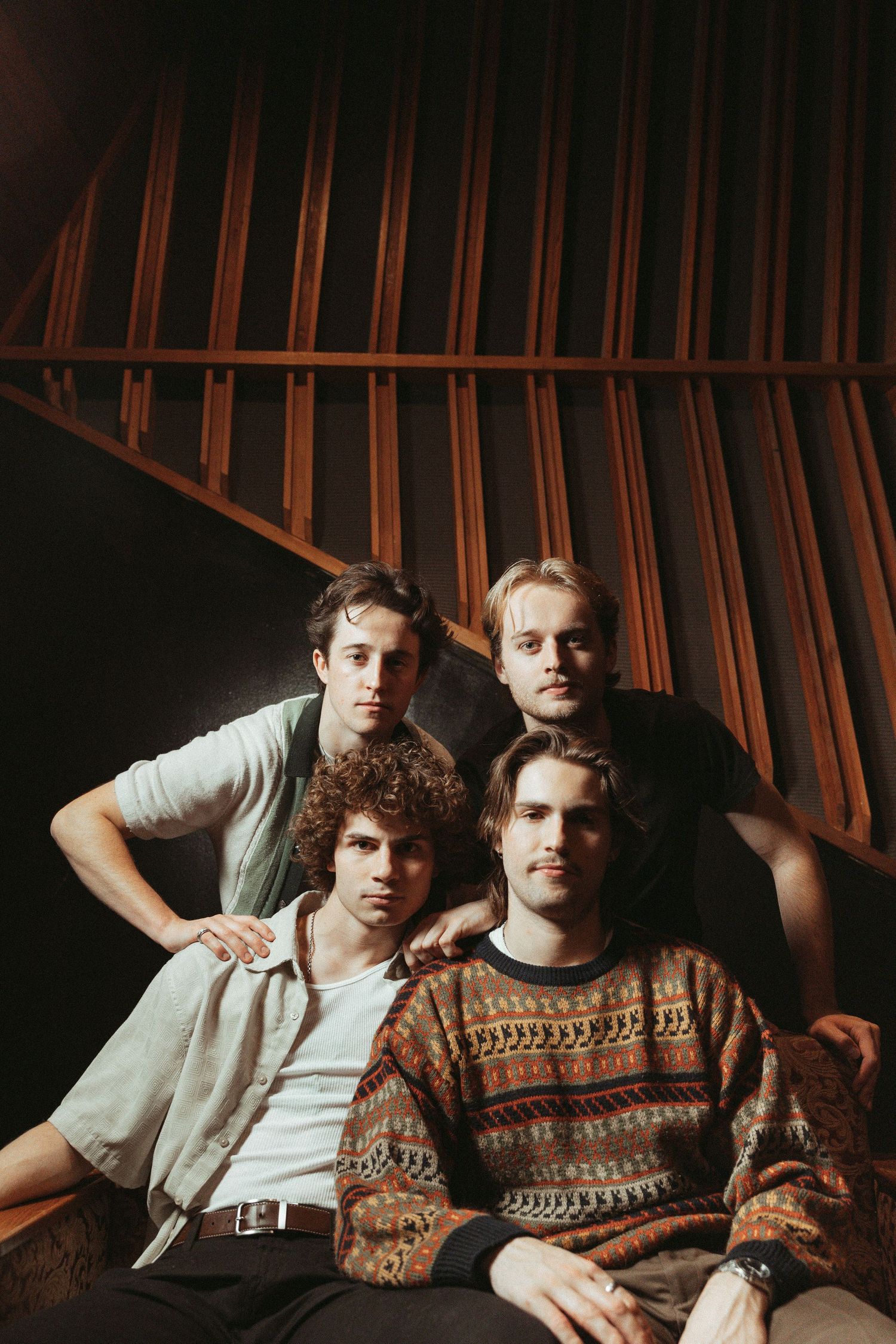 A group of four musicians pose together in vintage-style portrait with warm lighting and retro clothing against dark backdrop.