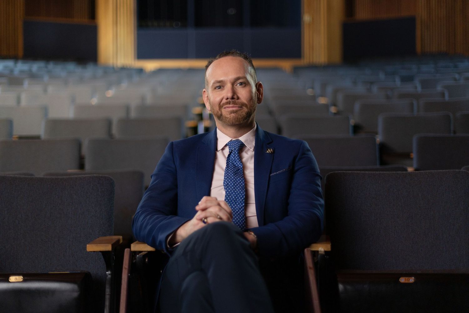 A person in a navy suit and blue tie sits confidently in a theatre-style auditorium with rows of empty seats behind them.