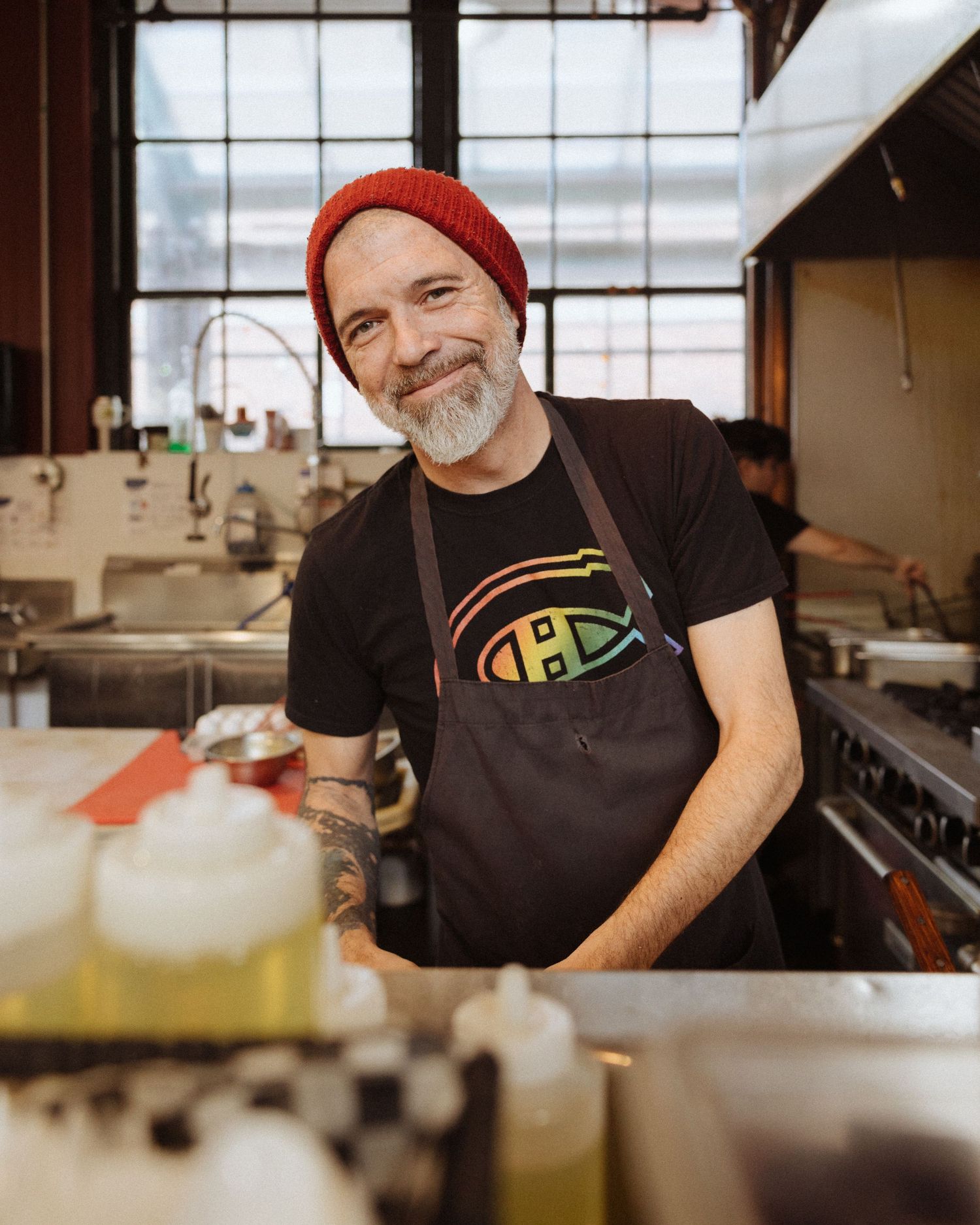 Someone wearing a red beanie and black apron smiles while working in a restaurant kitchen.