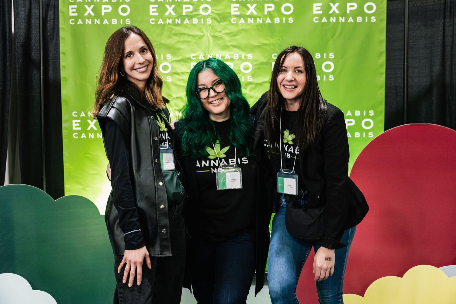 Three people wearing black attire pose together in front of a green expo banner with colorful decorative elements.