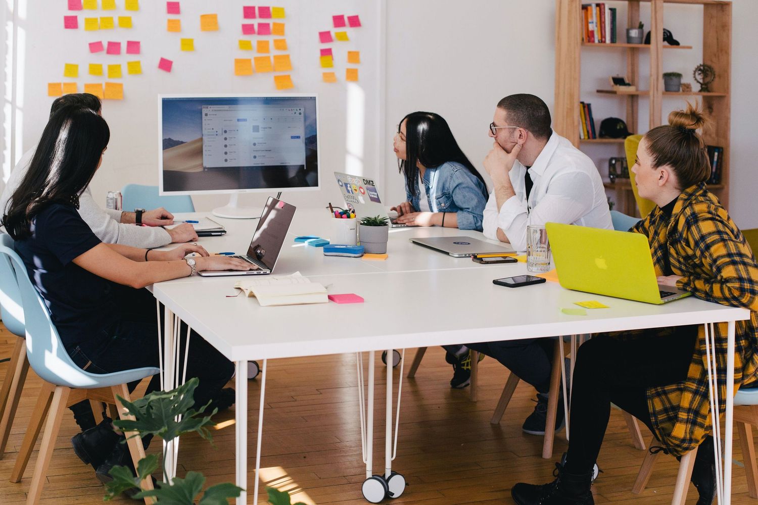 Team working together at white desk with colorful sticky notes on wall and laptops during creative office meeting.