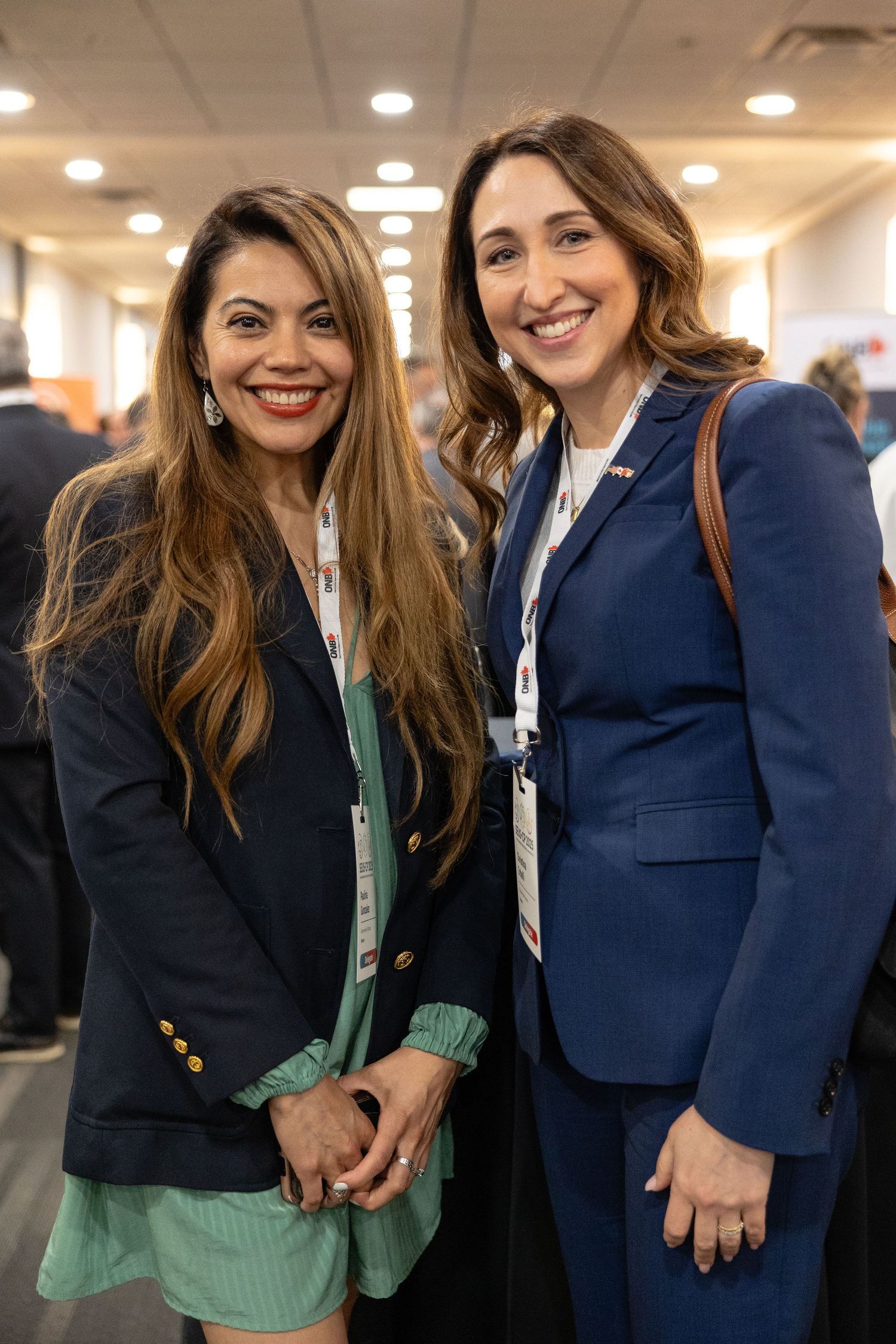 Professional colleagues in business attire smile together at a conference or networking event in a brightly lit indoor space.