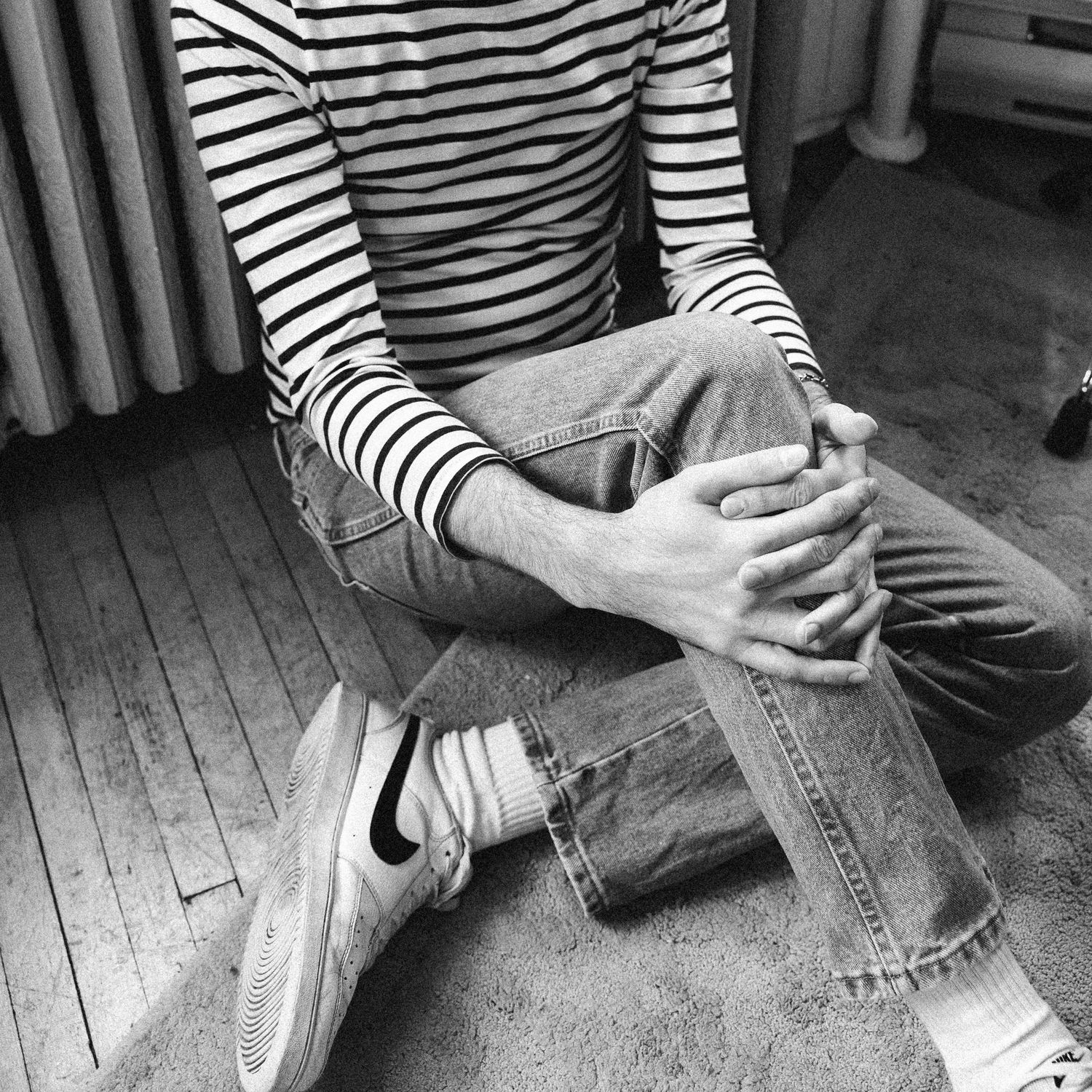 Person wearing striped shirt and jeans sits cross-legged on wooden floor with hands clasped over knee.