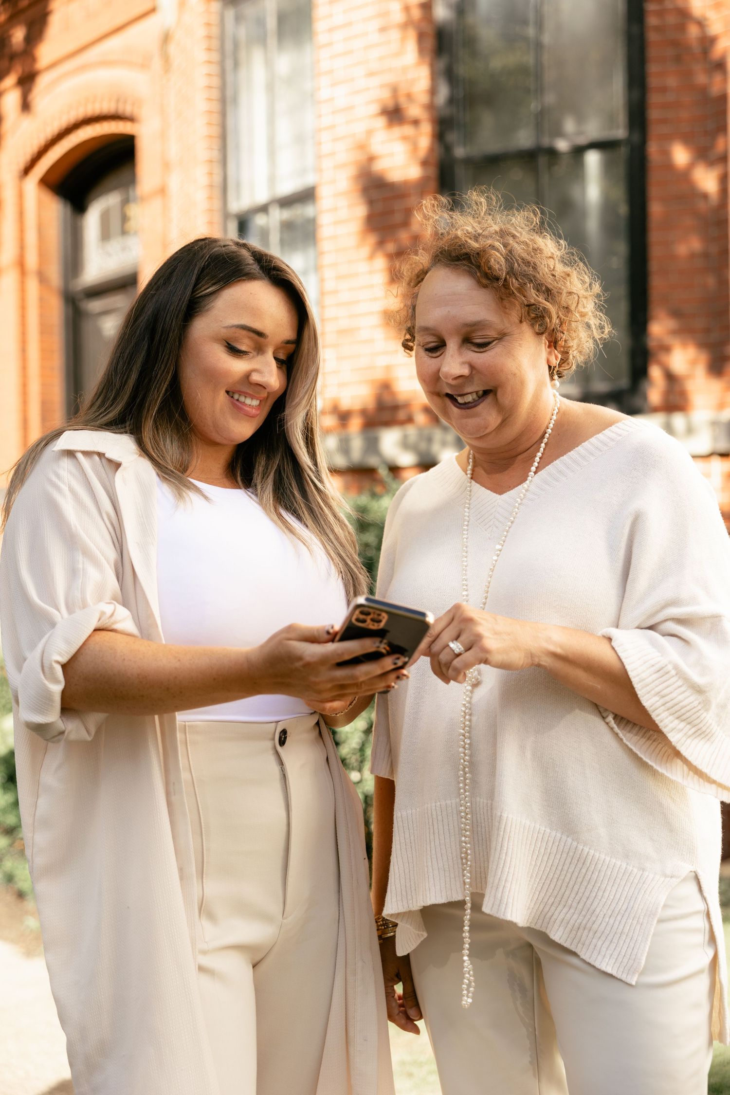 Two people in white outfits stand together looking at their mobile phones in an outdoor setting.