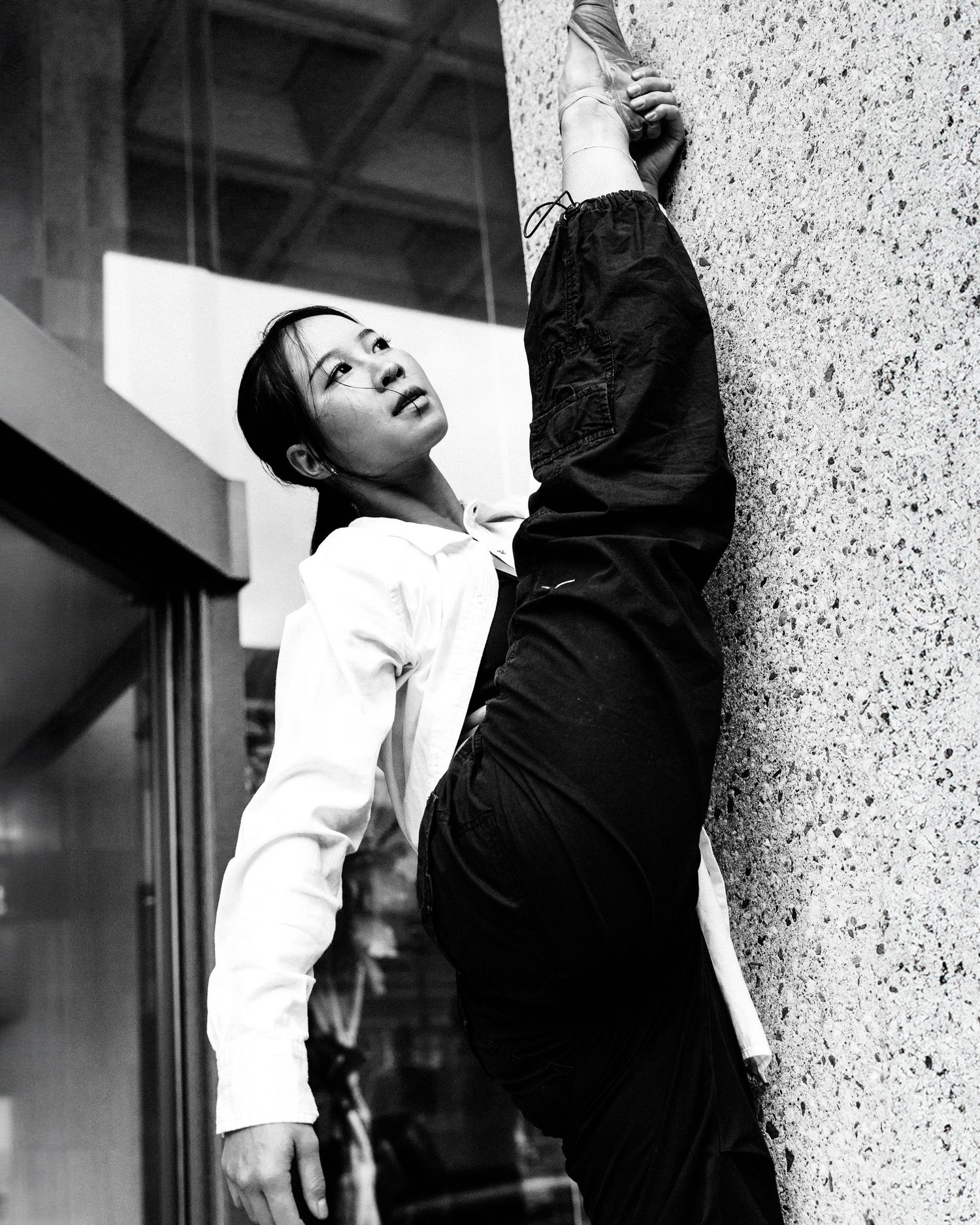 A dancer in white shirt and black pants performs an expressive contemporary dance move against a textured wall.