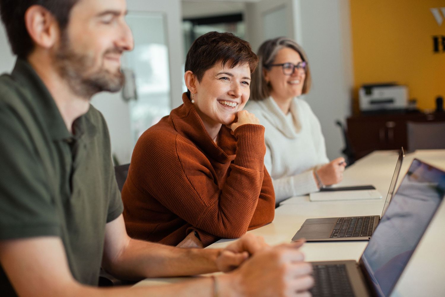 Three people sitting at a desk smile while working together on laptops in a bright modern office setting.