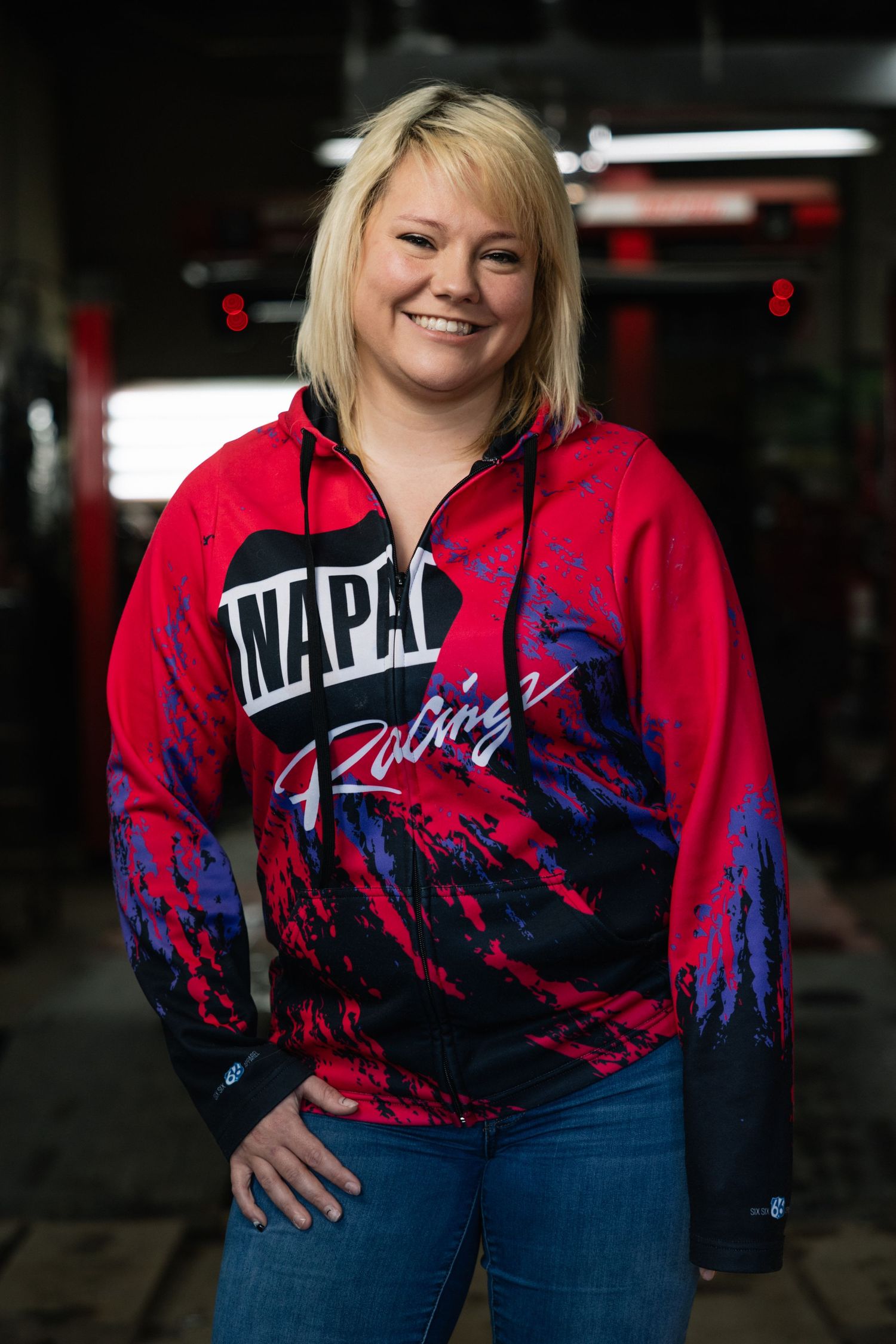 Person wearing a red and blue NAPA Racing sweatshirt with paint splash design and jeans in a garage setting.