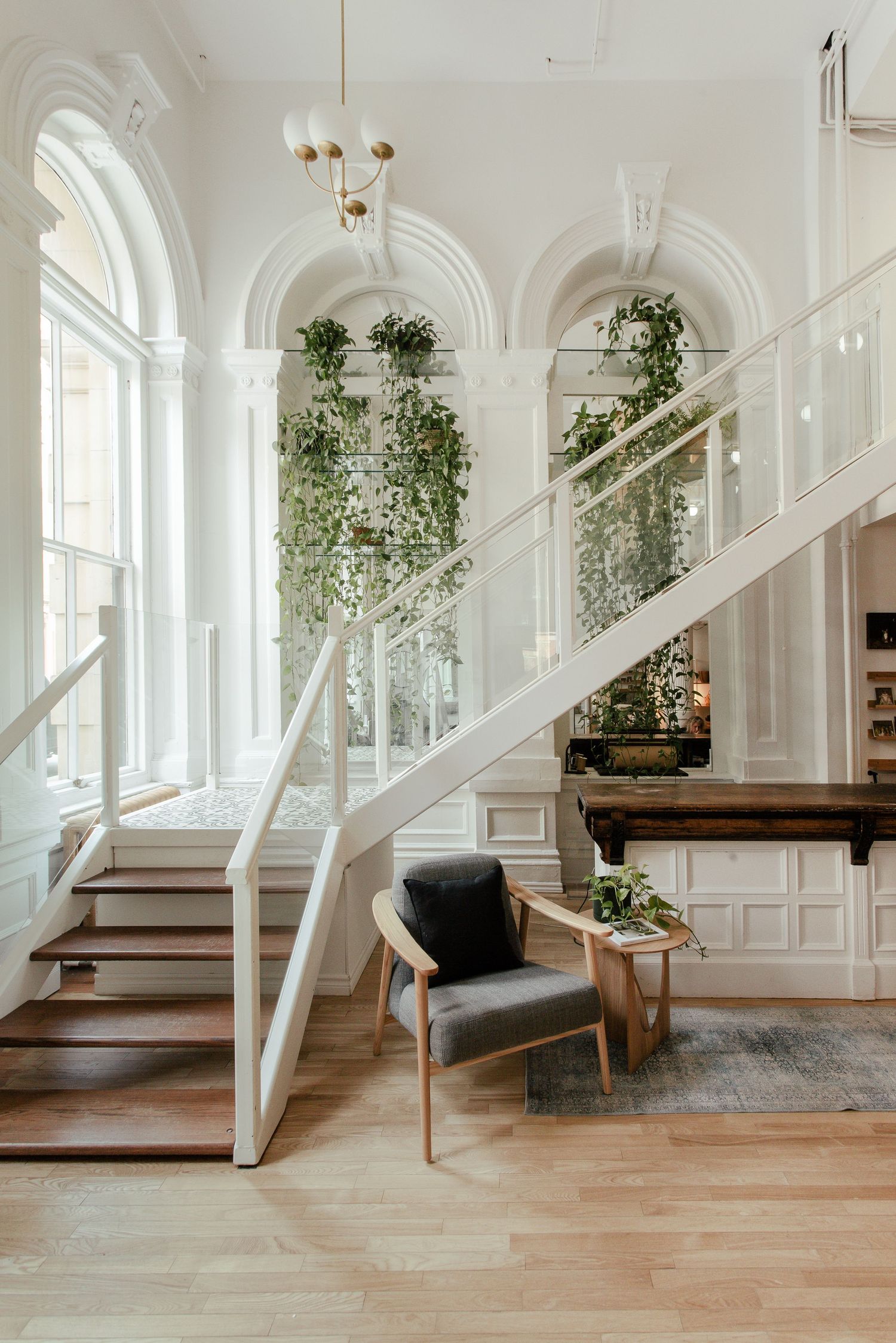 Bright interior hallway with white paneled walls, wooden floors, hanging plants, and modern grey accent chairs in sequence.
