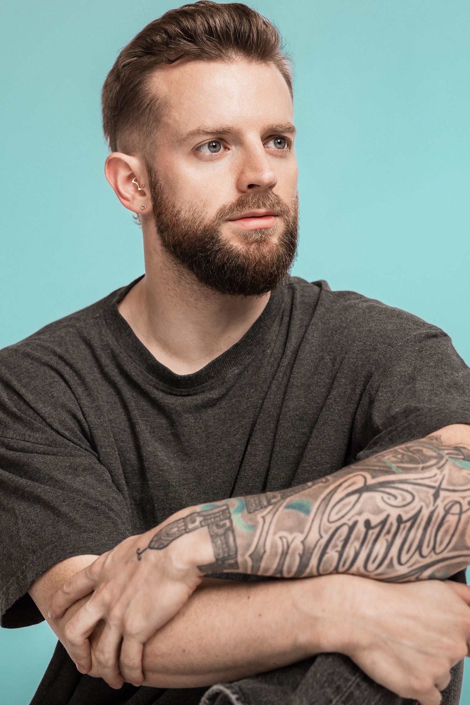 Studio portrait of a bearded individual in a dark t-shirt with a tattoo sleeve against a turquoise background.