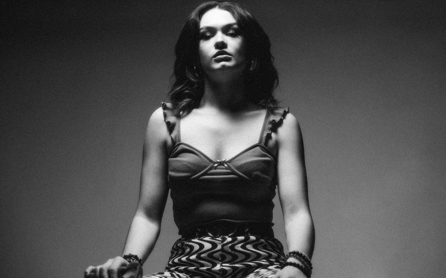 Black and white portrait of a person wearing a bustier top and patterned skirt while sitting against a gradient background.