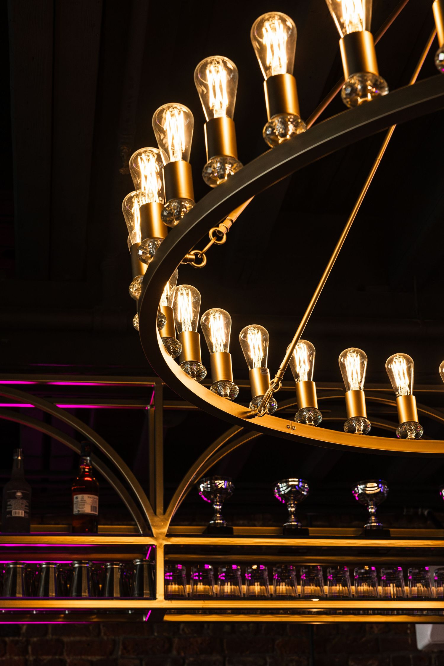 Elegant curved lighting fixture with vintage Edison bulbs illuminates a bar with wine glasses and bottles on display.