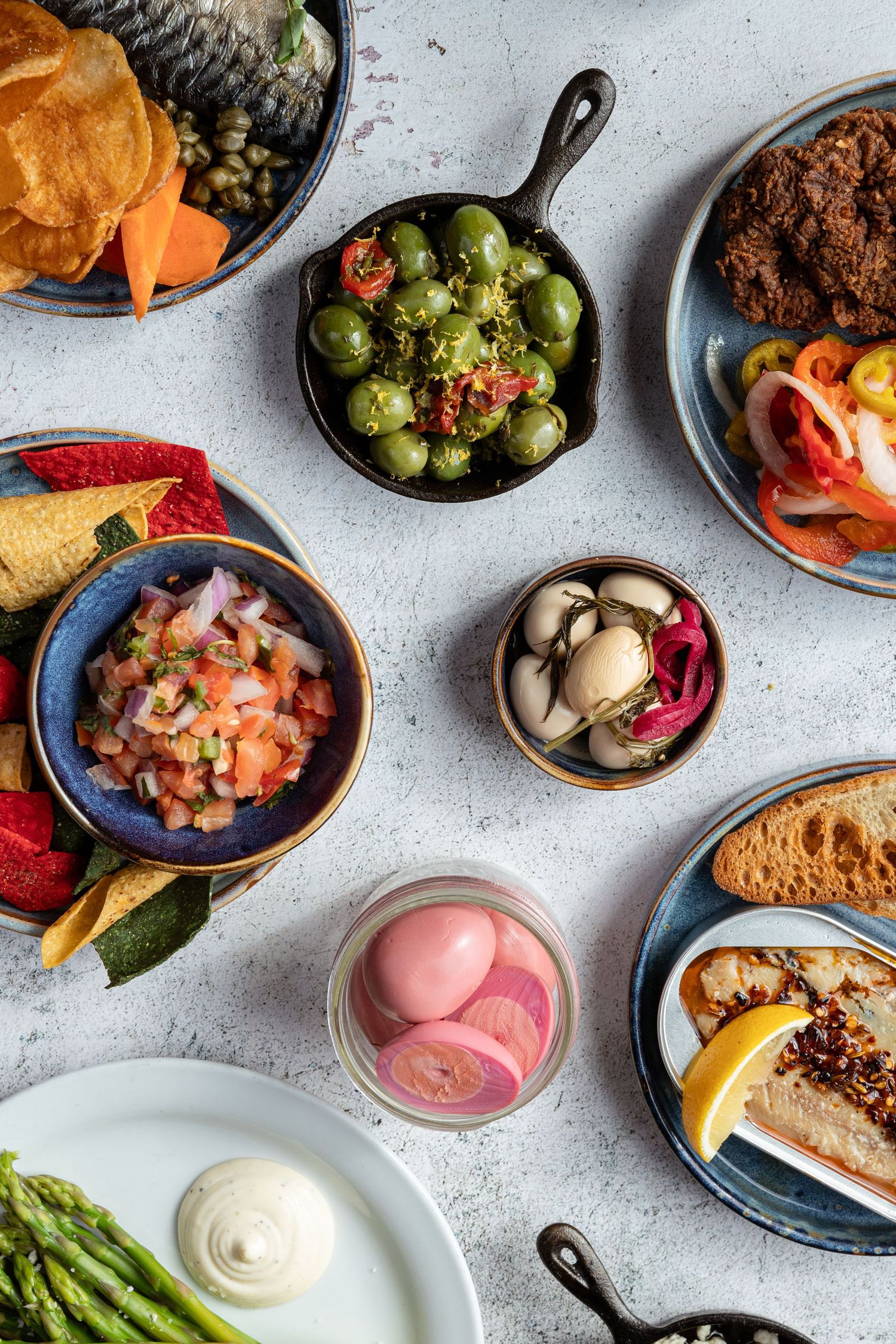 Overhead view of colorful tapas dishes including olives, pickled vegetables, seafood, asparagus and bread on a light gray surface.