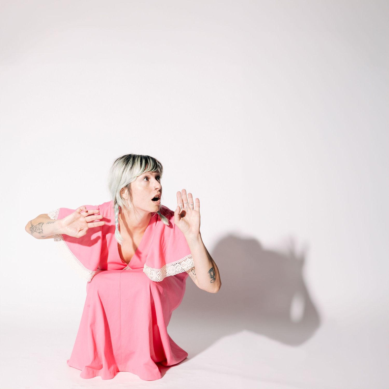 Figure in a pink ruffled dress crouches against white background while shouting with cupped hands.